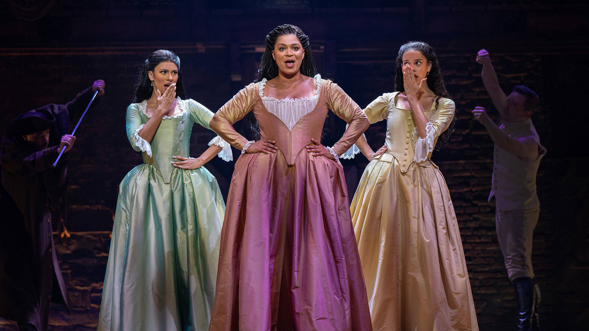 Three women with medium to dark complexion wearing pastel colored colonial dresses with large skirts and white sleeve ruffles stand together. The woman at the center has her hands on her hips while singing, the other two are slight behind each with a hand to their mouth in surprise.