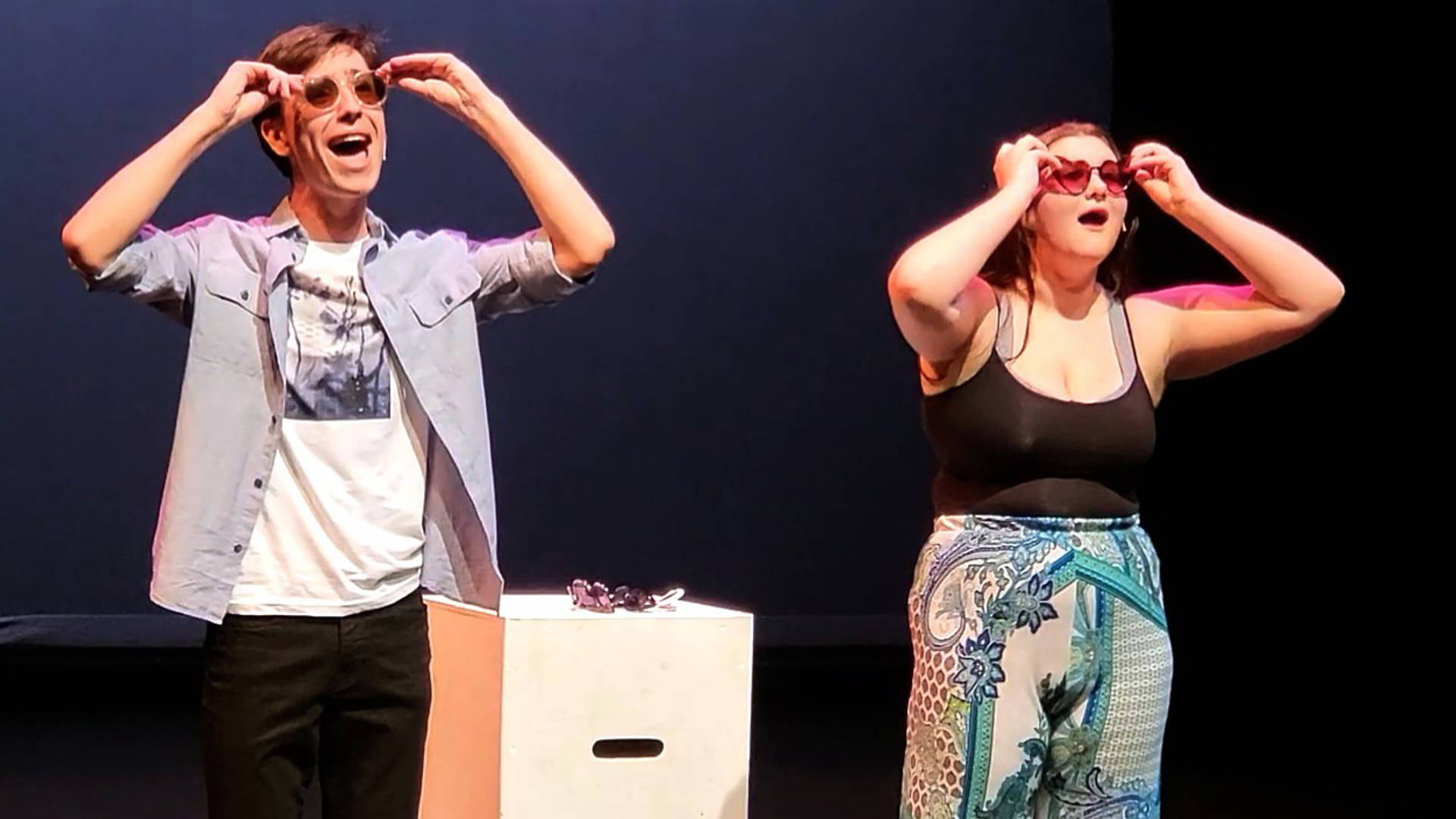 Two high school students performing theater. They are wearing warm weather clothing and sunglasses.