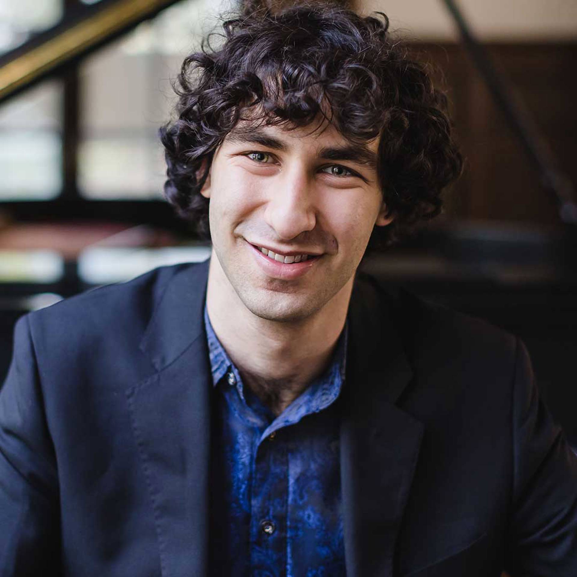 Headshot of a young caucasian man with medium length curly dark hair and greyish eyes. He is smiling and has a dark jacket on over a blue shirt.