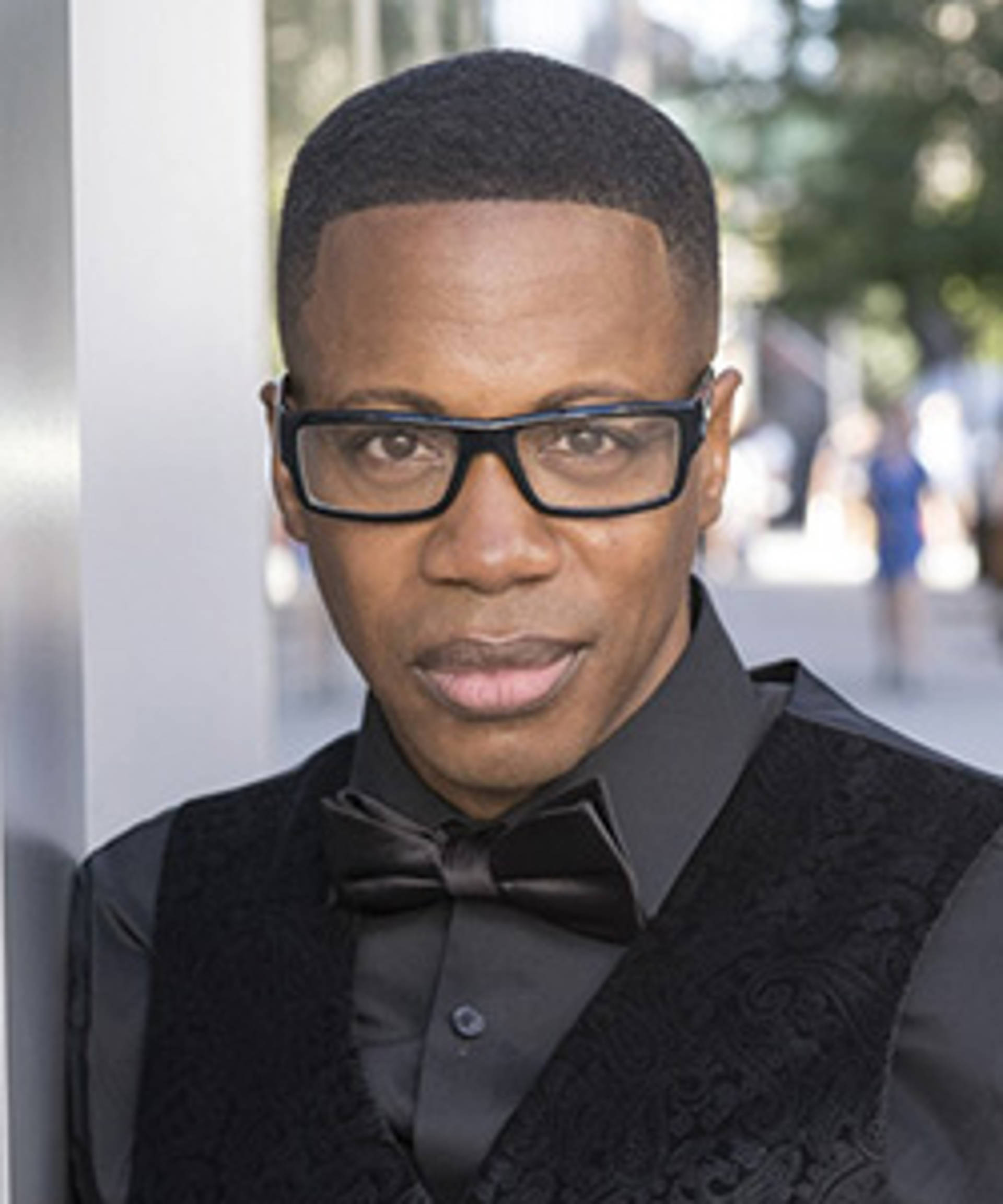 A young african american man with short hair, glasses and a dark shirt, vest, and black bowtie