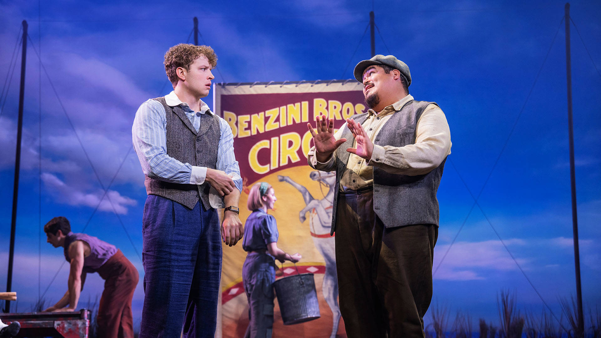 Two men with short brown hair have an animated conversation in front of a circus sign.