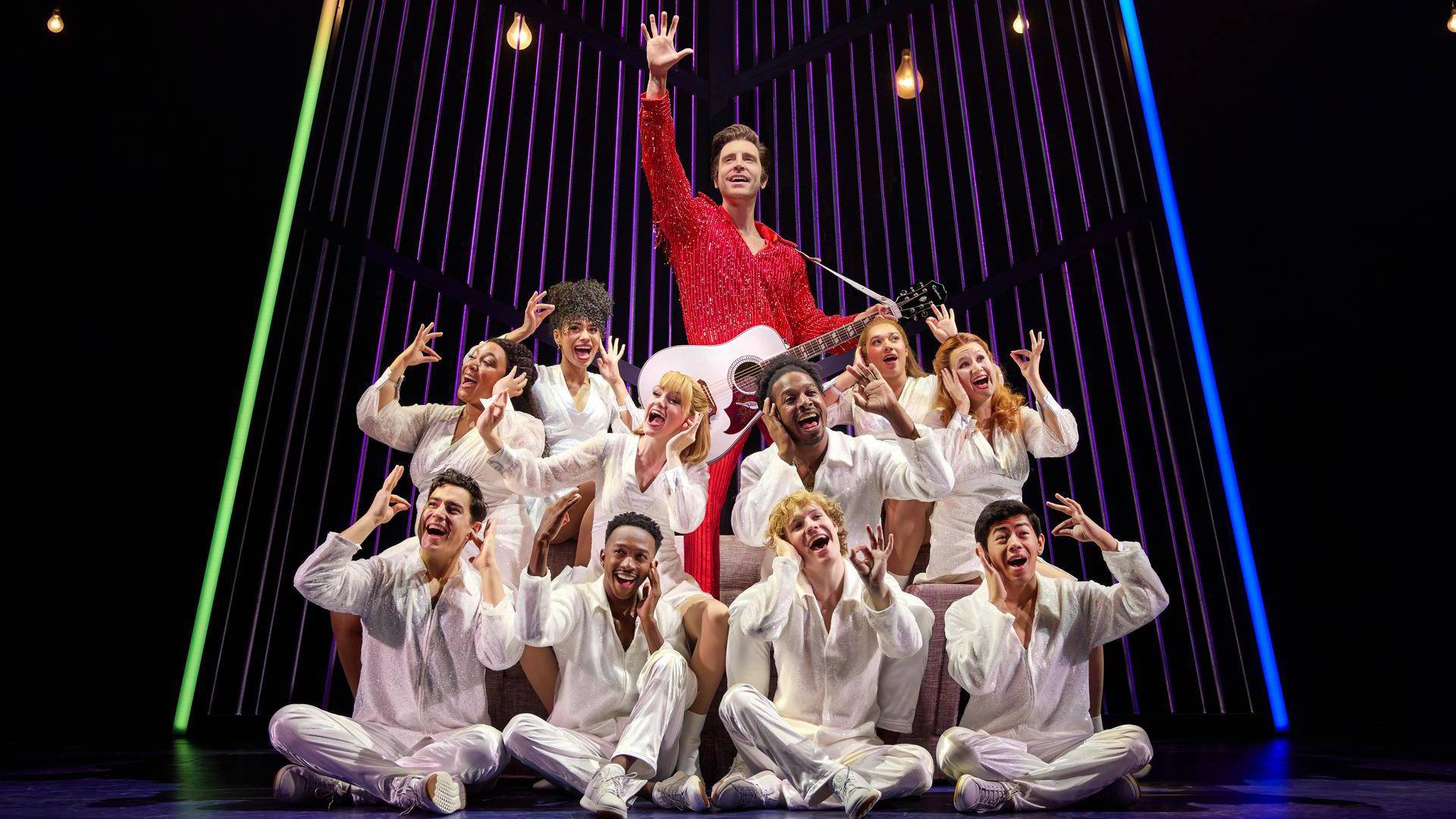 A man with light complexion and short greased brown hair in a bright red outfit with a white guitar. A group of people in all white outfits are sitting on the ground in front of him.