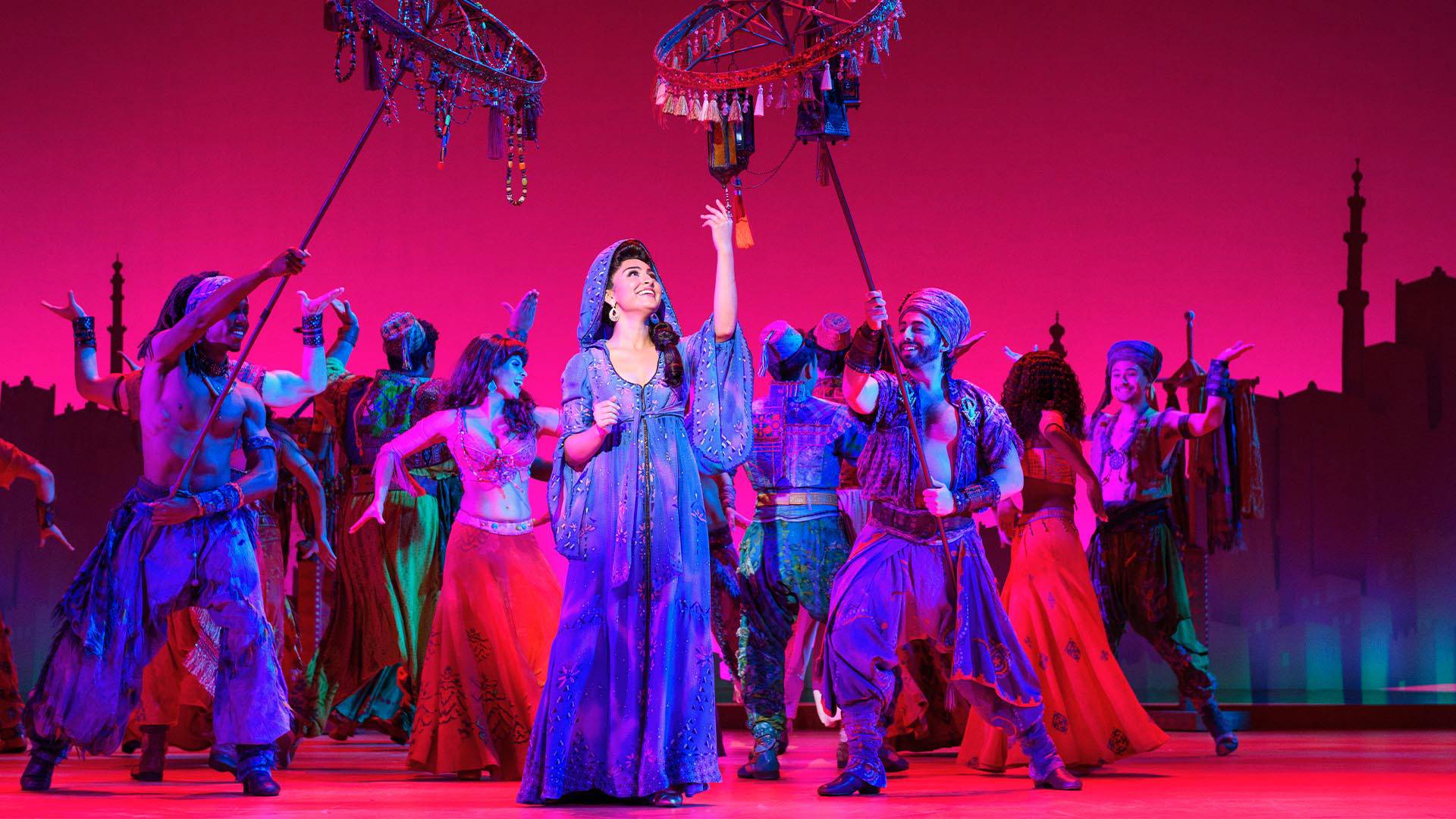 A woman standing in front of many dancers. Two men hold decorative rings of jewelry above her head. Everyone is dressed in Middle Eastern clothing. They stand in front of a pink backdrop.