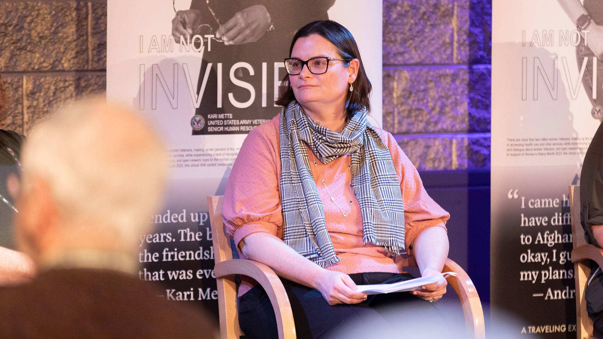 A woman with light complexion, dark medium hair and a scarf sitting in a chair and holding a piece of paper in her hands.