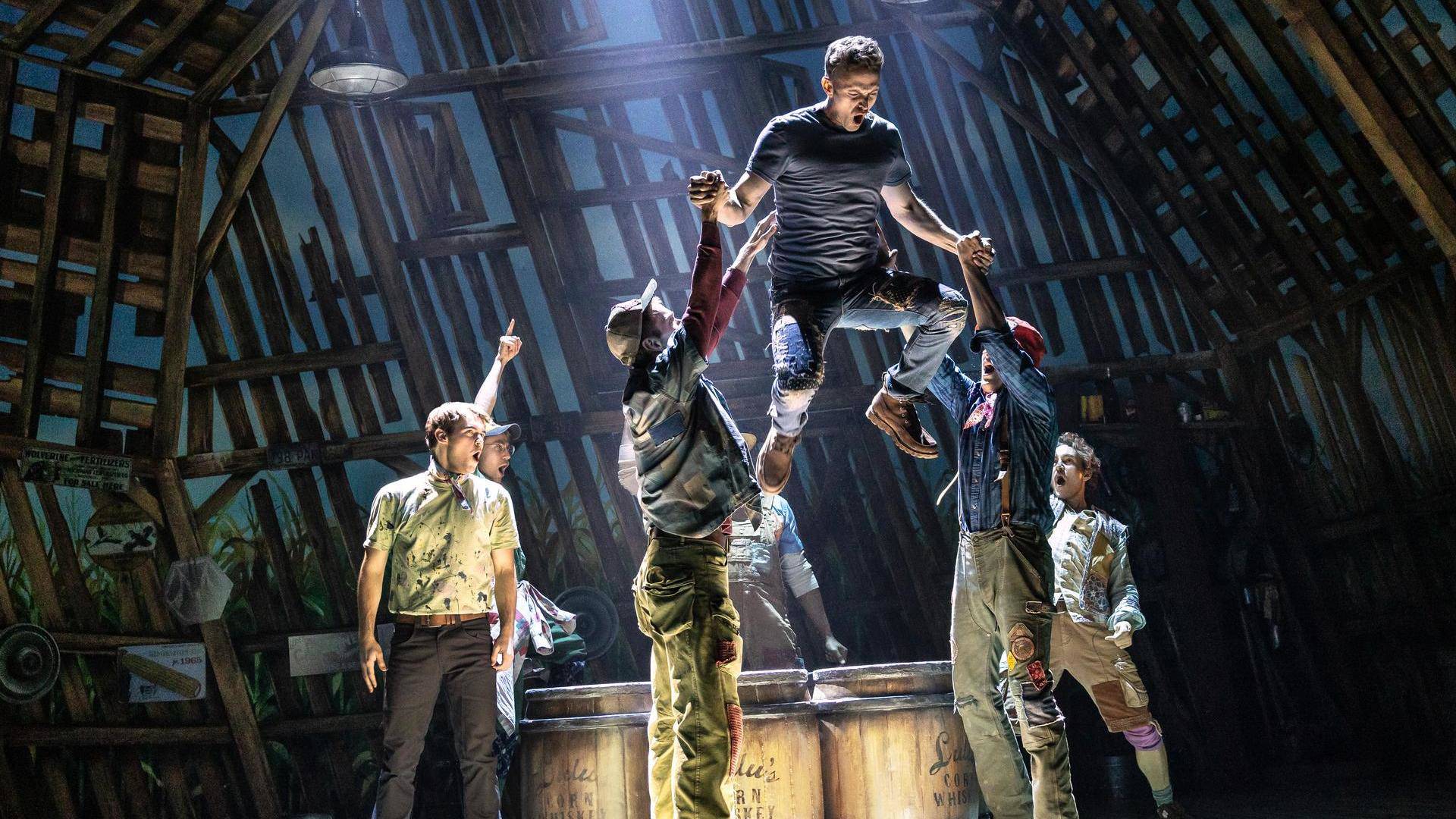 a man in a grey tshirt and jeans leaps of a group of barrels, helped by other men singing around him. the lighting is dark.