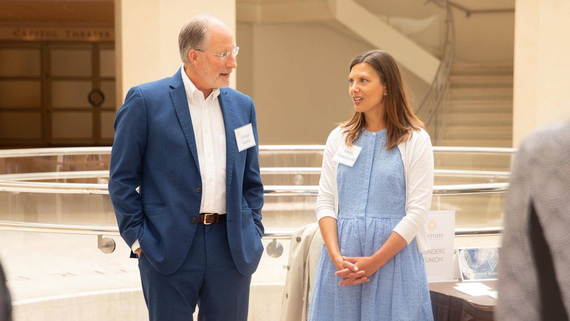 A man and a woman stand talking in a Overture's Rotunda. The man has light complexion grey short hair and a blue suit. The woman has light complexion long brown hair and a blue dress.