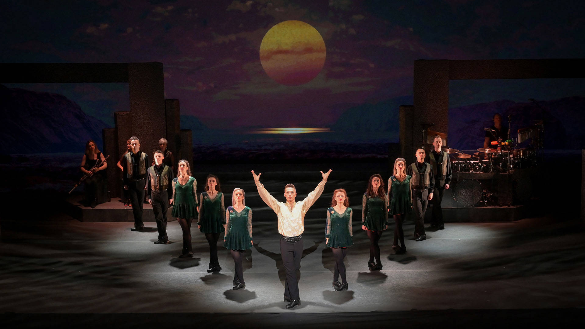 Irish dancers in dark green velvet costumes, stand in a triangle formation against a lowlit sunset backdrop. At the center and tip is a light complexion dancer with short blonde hair wearing a white blouse and black pants, they have both hands reaching upwards.