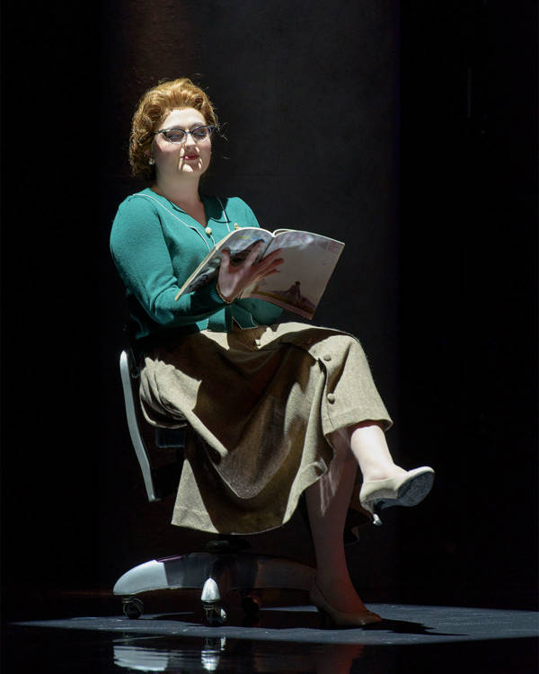 A caucasian woamn in midcenury clothing stting in a chair reading a magazine.