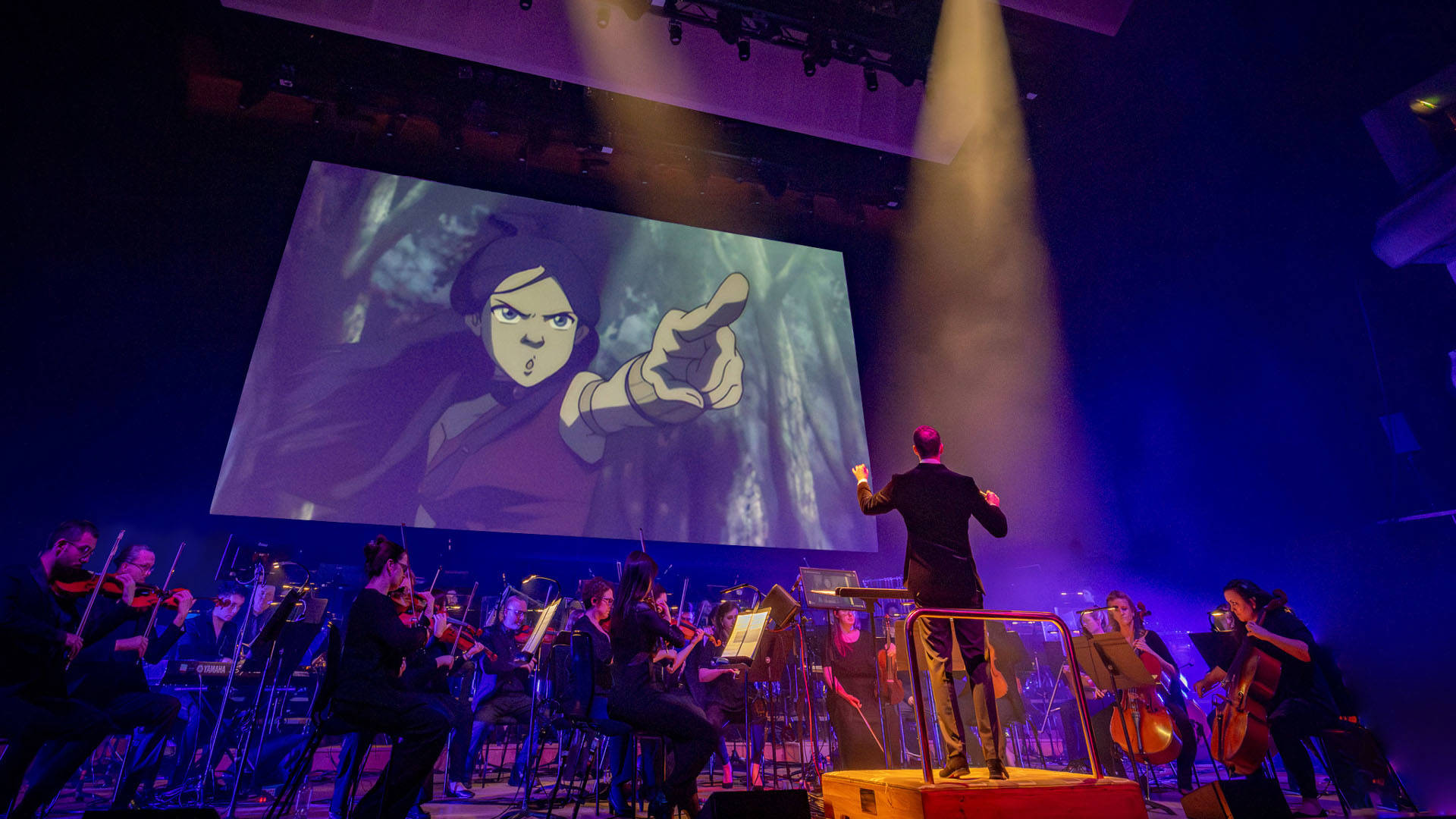 An orchestra performing on stage with a standing composer. A screen plays a scene from Avatar: The Last Airbender behind them, showing a young girl with long brown hair pointing at the distance.