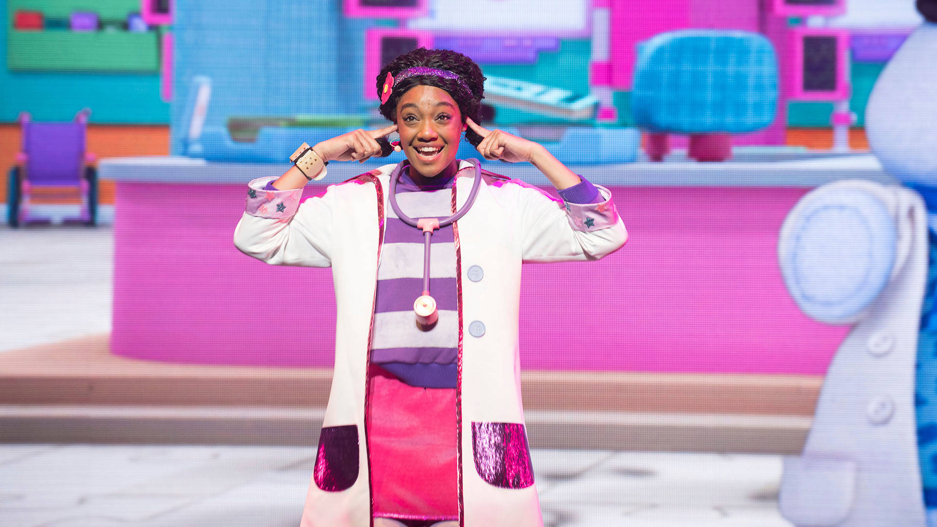 A young African American woman smiles and points both index fingers towards her head. She is wearing a striped purple and white shirt with a doctors coat and stethoscope around her neck.