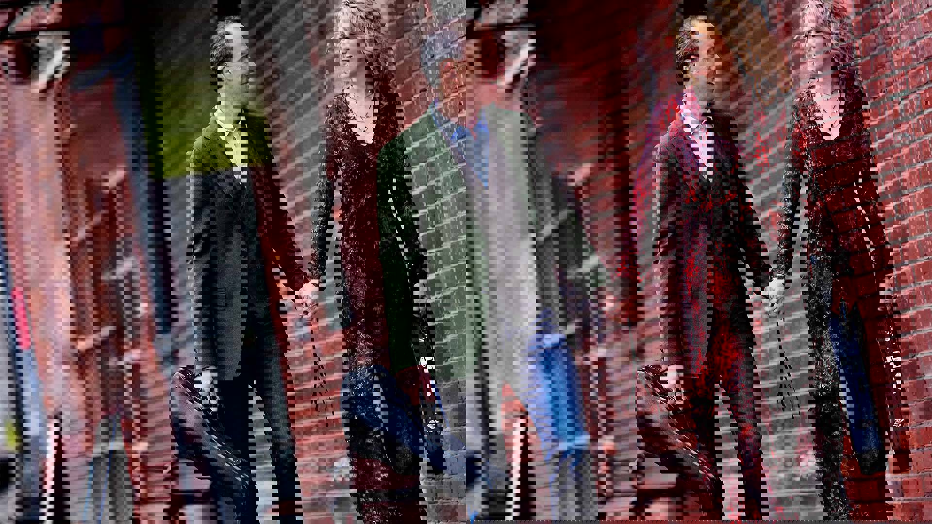 A caucasian man and woman walk together holding hands and carrying violin cases. The man has short graying hair and a green jacket with jeans. The woman has long blonde hair and a red and black dress on.