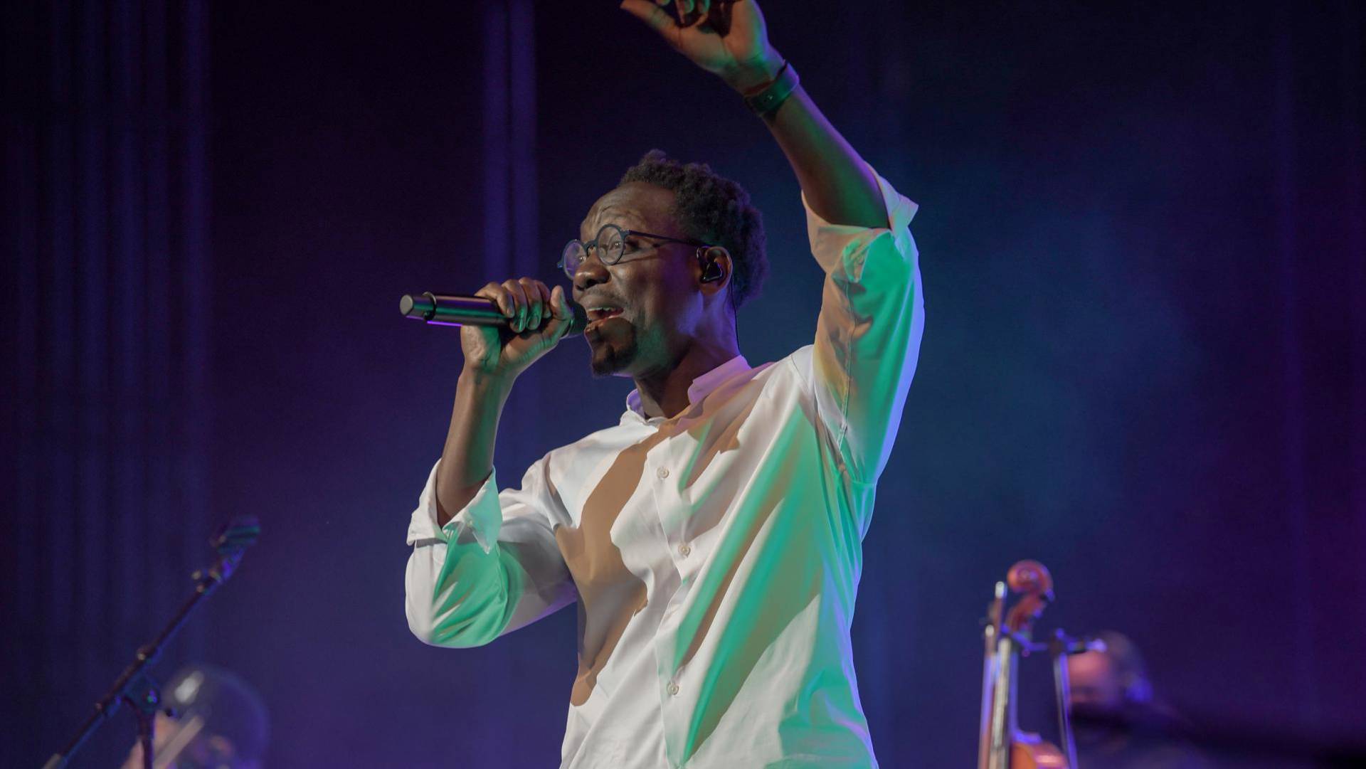 A man with dark complexion, a white shirt, and a short hair speaks into a microphone on stage.