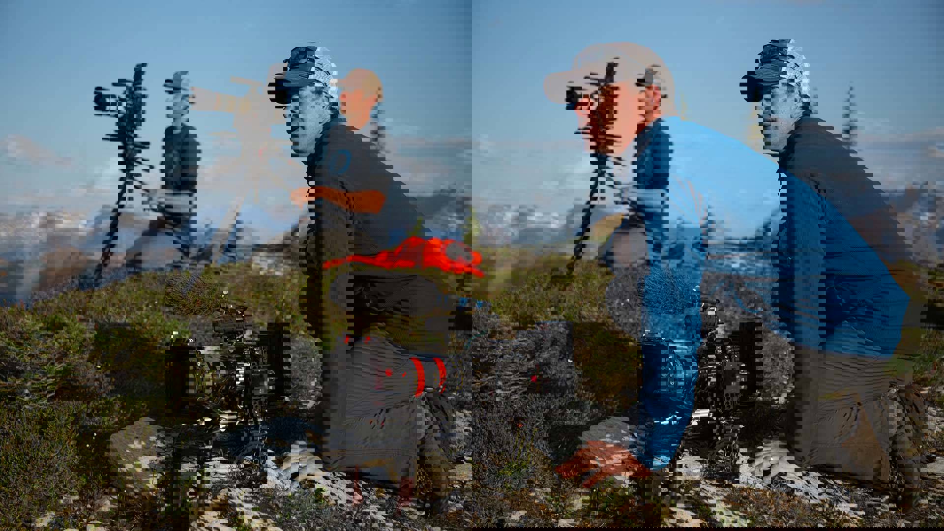 Two caucasian men holding video cameras crouched on a grassy hill backed by mountainous terrain.