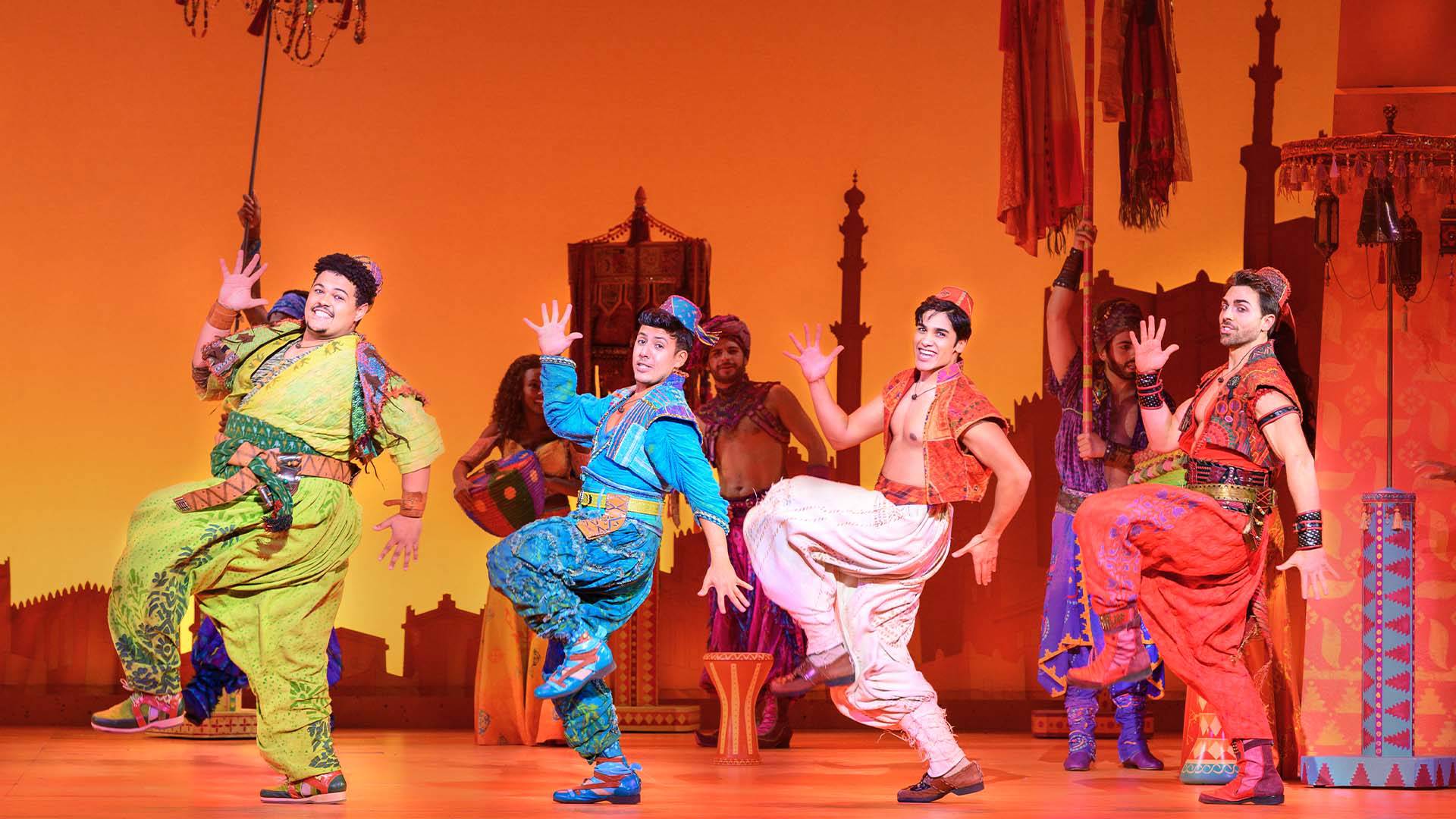 Four men dancing on a stage with an orange, backdrop. They are wearing Middle Eastern style clothing. They are dancing in a line, with their right arms and left legs in the air.