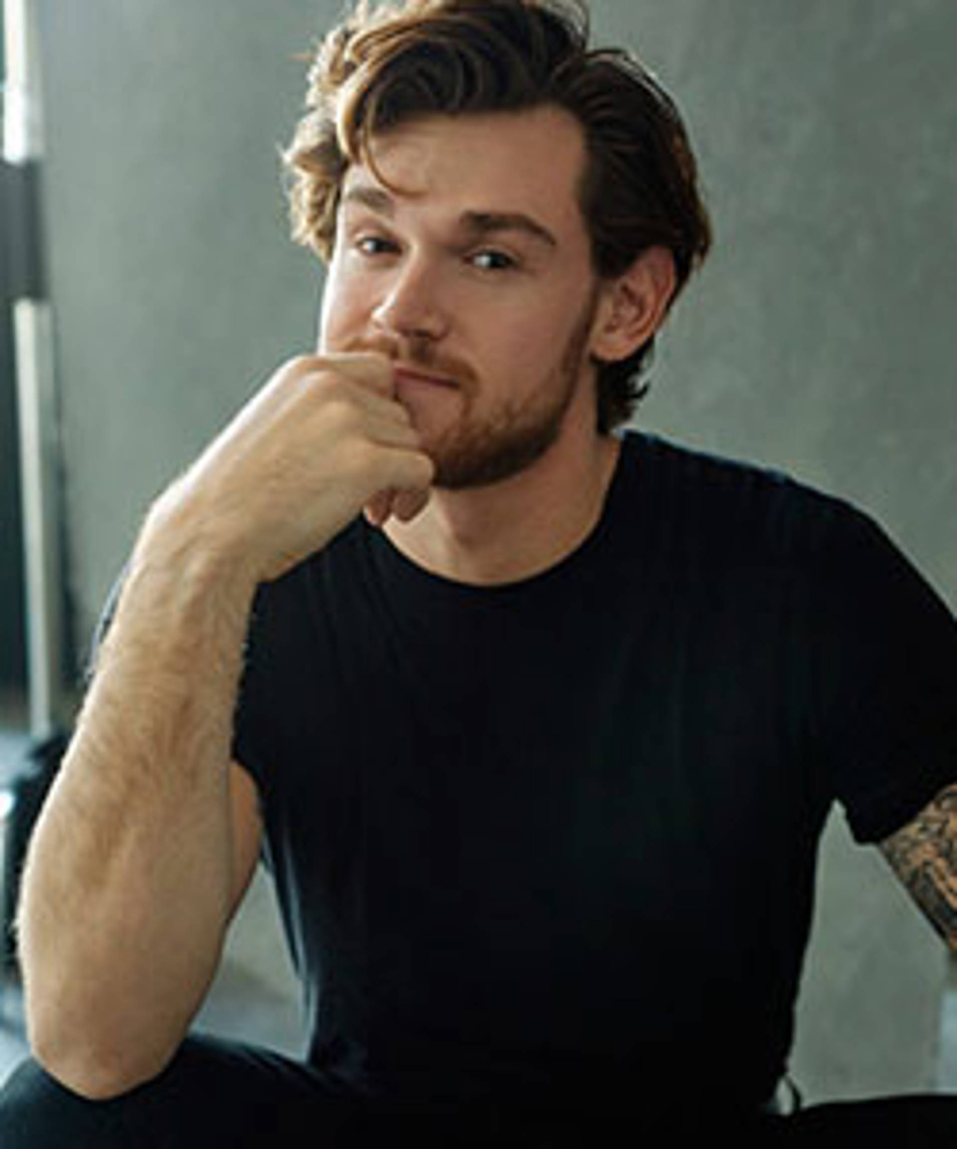 Headshot of a caucasian man with dark eyes and wavy brown hair. He is sitting and resting his head against his hand
