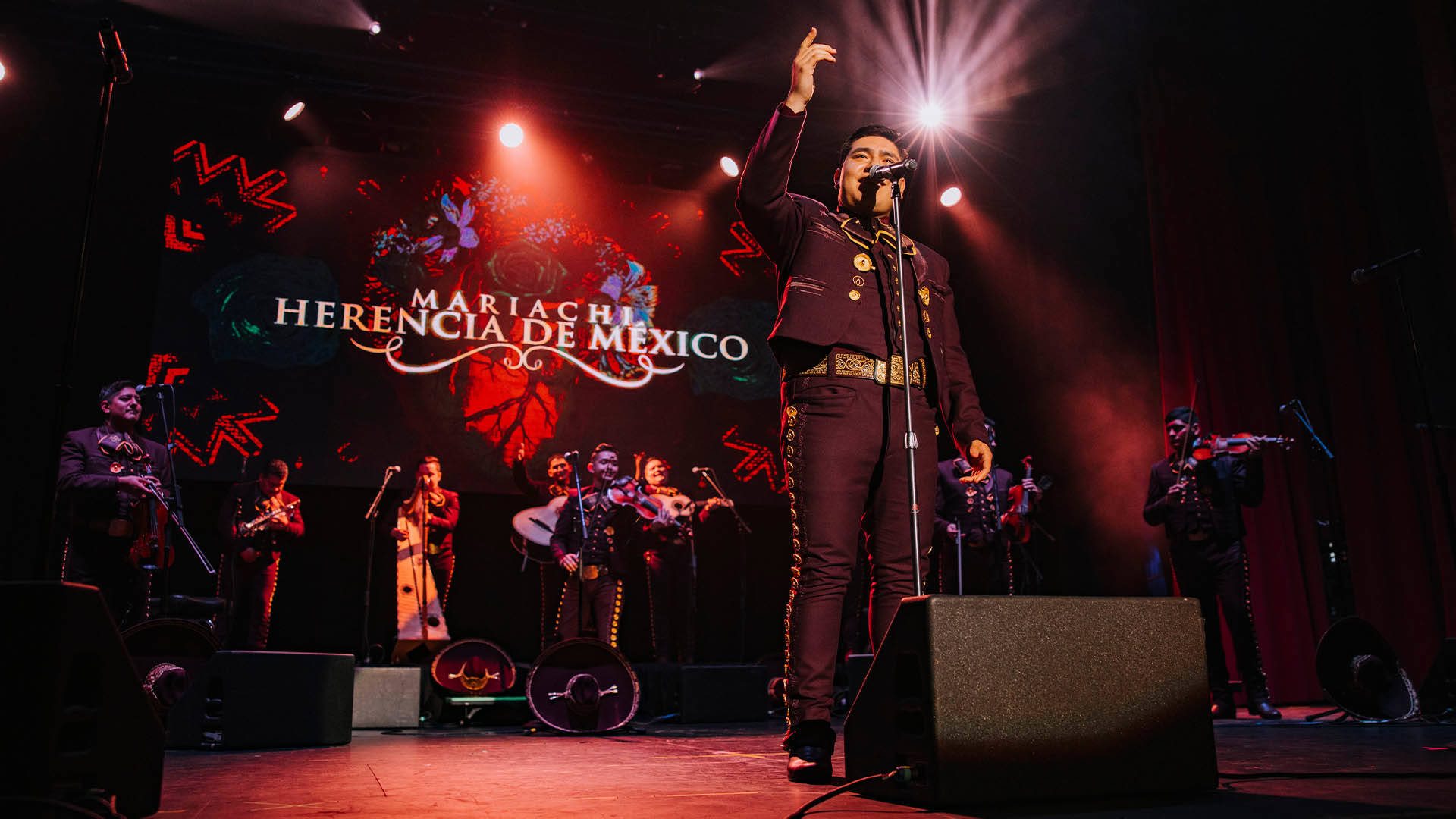 A person in a dark, Mariachi outfit. They are standing in front of a microphone on a stage. Behind them are additional band members and a sign that reads "Mariachi Herencia de Mexico".