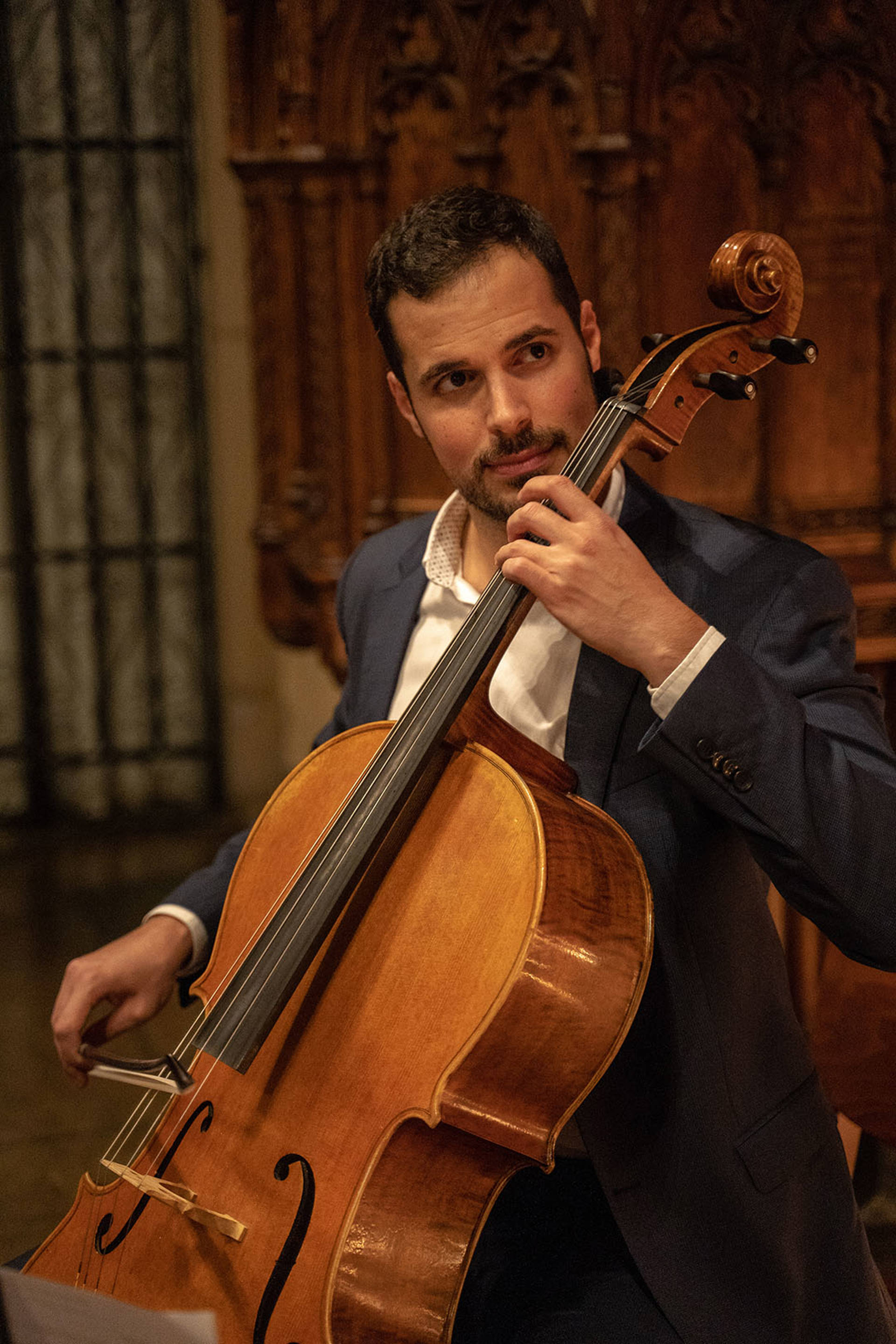A man with light complexion, dark hair and a light beard in a blue suit coat playing a cello.