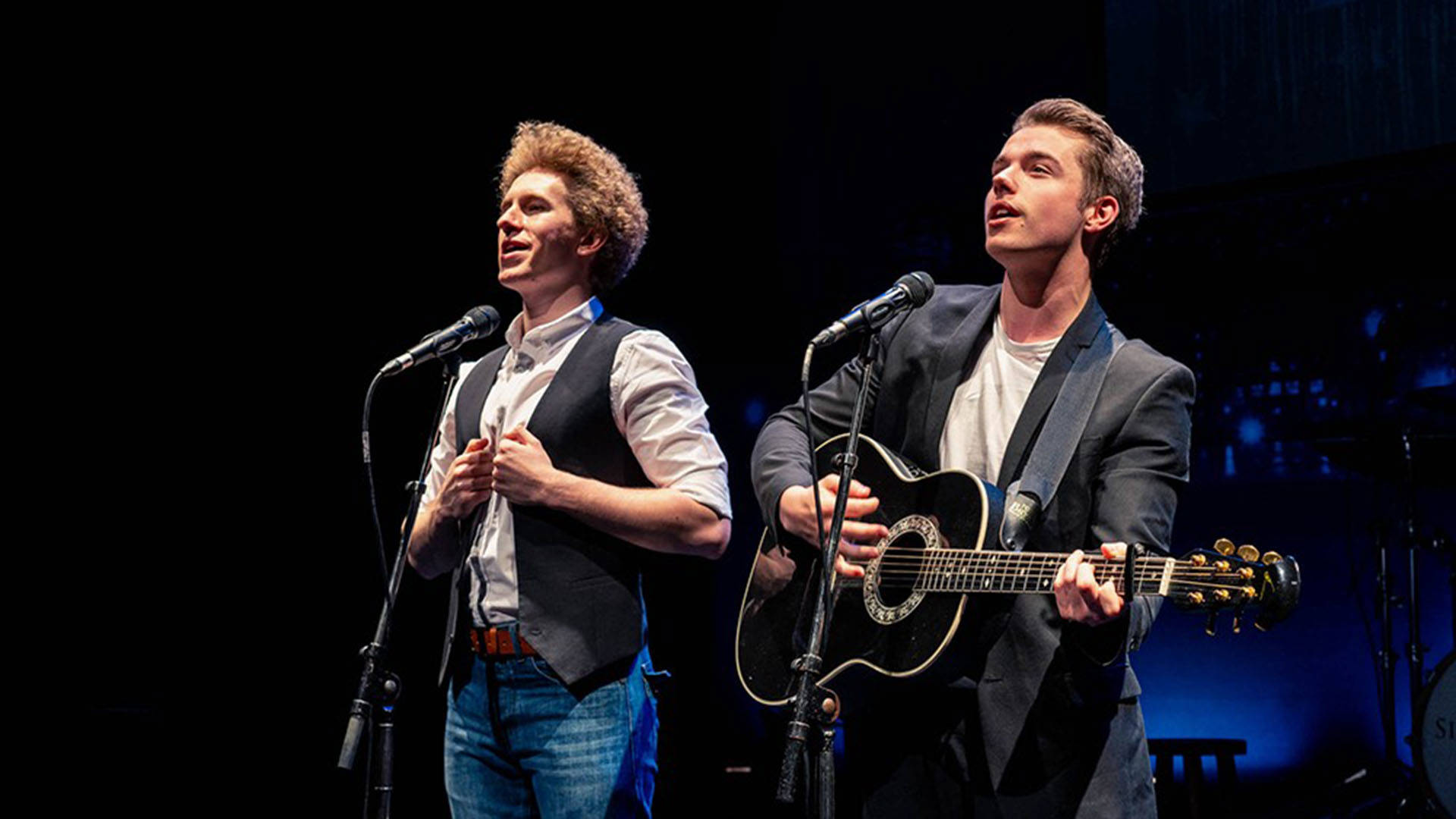 Two white men standing behind microphones. The one on the left has light, curly hair and is wearing a grey vest and white shirt. The one on the right has short, brown hair, is wearing a grey jacket and playing a black guitar.