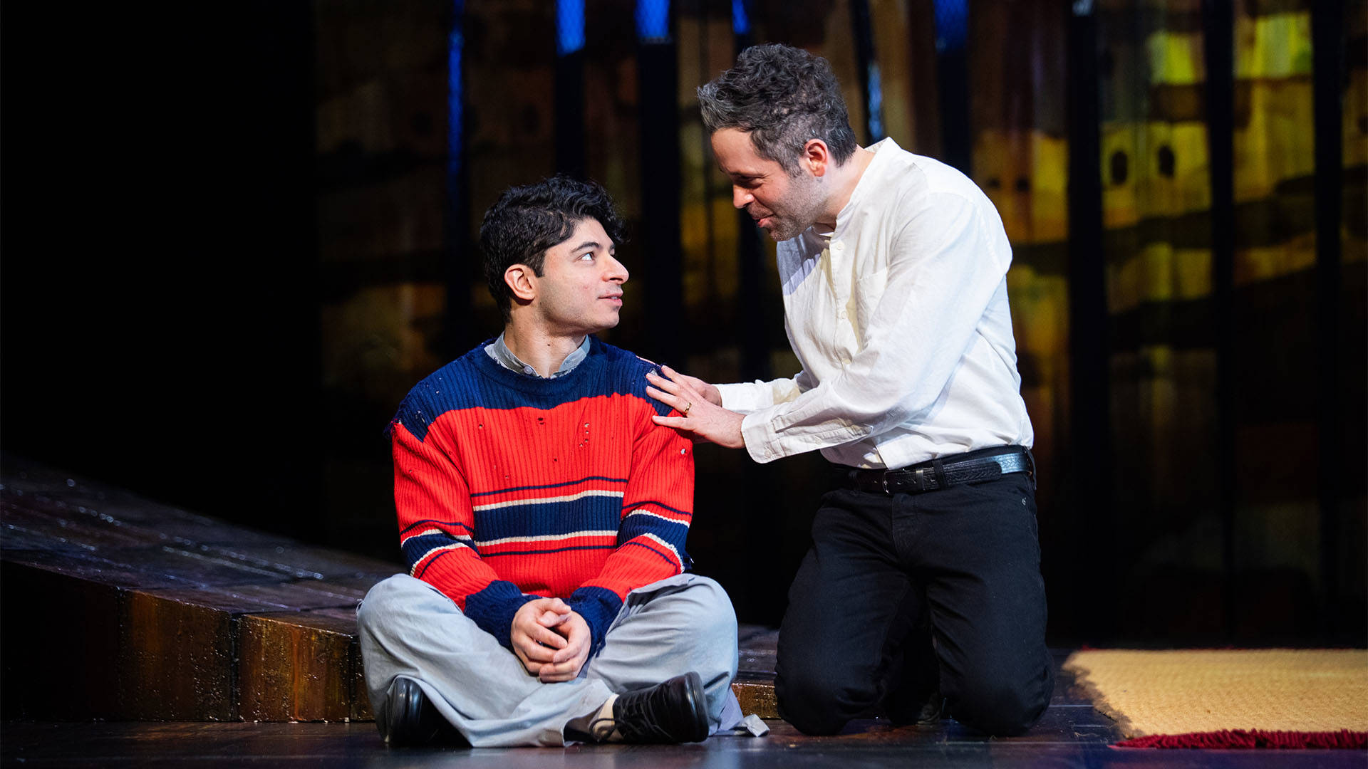 A young person with medium skin tone and dark short hair sits cross-legged with arms in their lap. They are wearing a red and blue striped sweater and light blue pants. A middle aged man with light skin tone wearing a white button up shirt sits next to them with his hands on their shoulder in conversation.