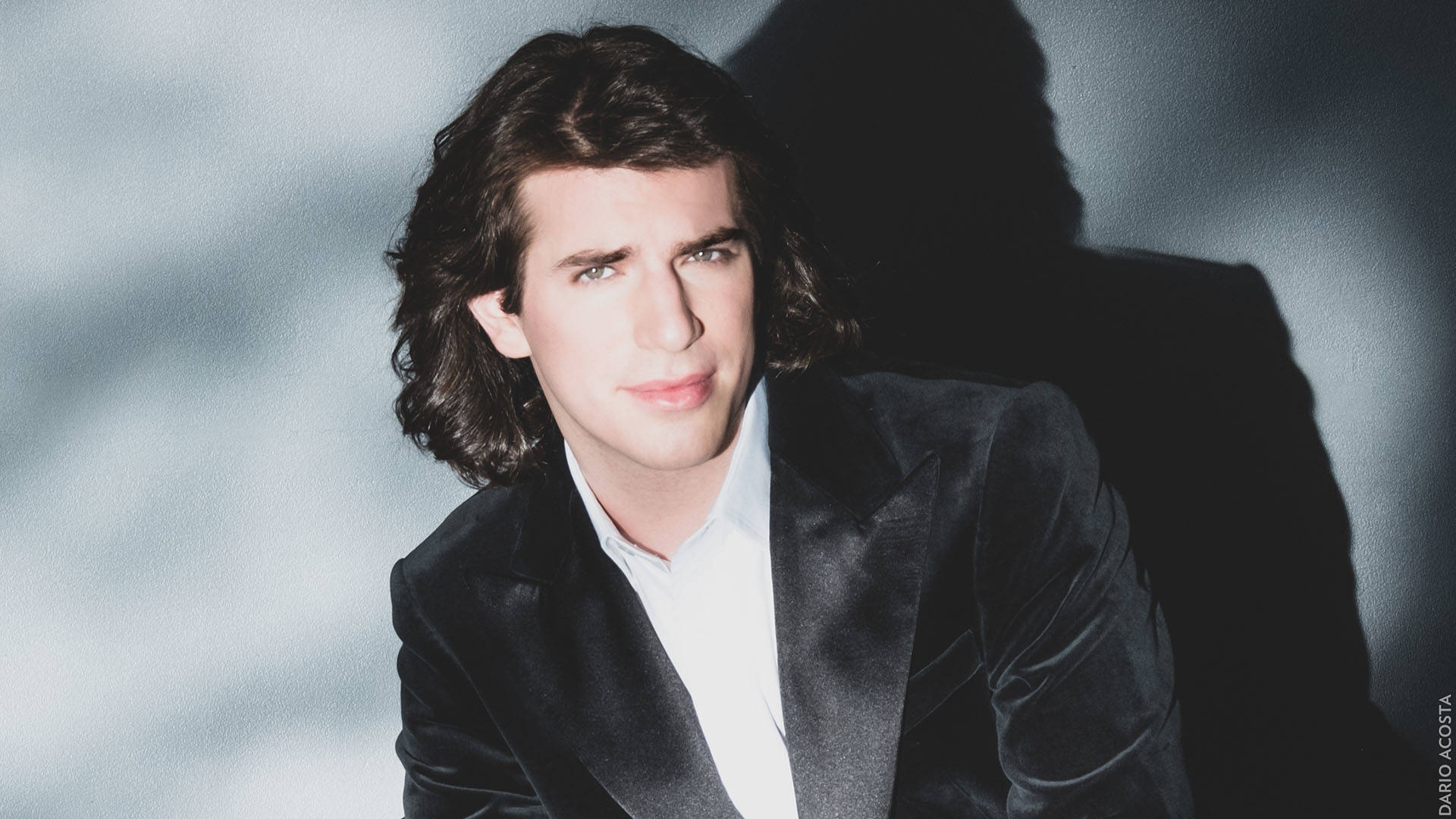Headshot of Llewellyn, a young man with light complexion and shoulder-length wavy dark brown hair, wearing a black velvet suit jacked and a white collared shirt against a grey backdrop.