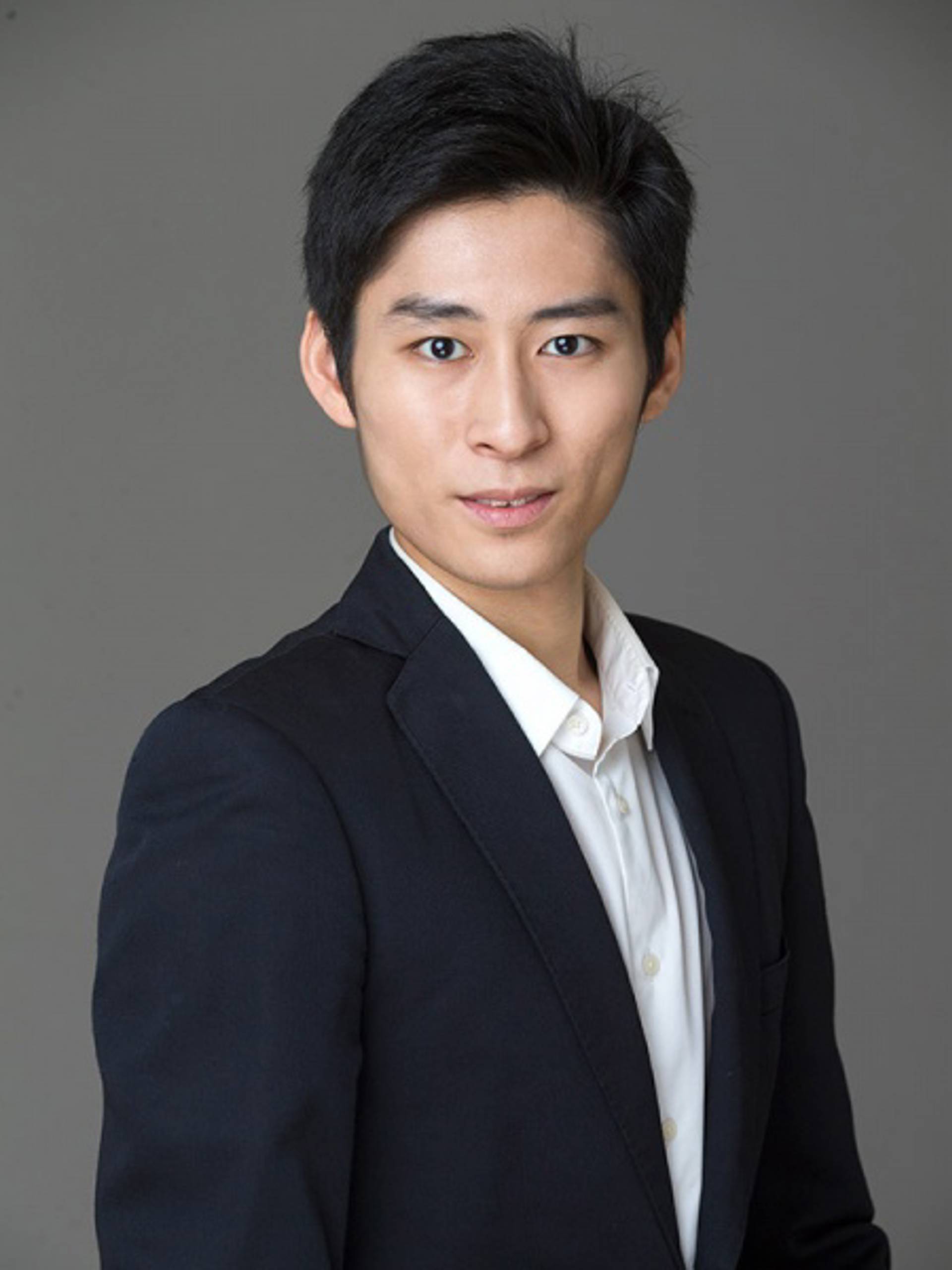 Headshot of a young man with medium complexion, short black hair, a white collared shirt and a dark jacket against a grey background