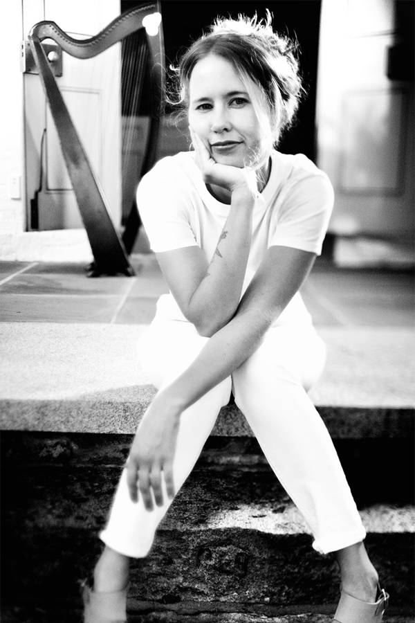 Grayscale image of a caucasian woman sitting and resting her head on her hand. She has a white outfit on. There is a harp standing behind her.