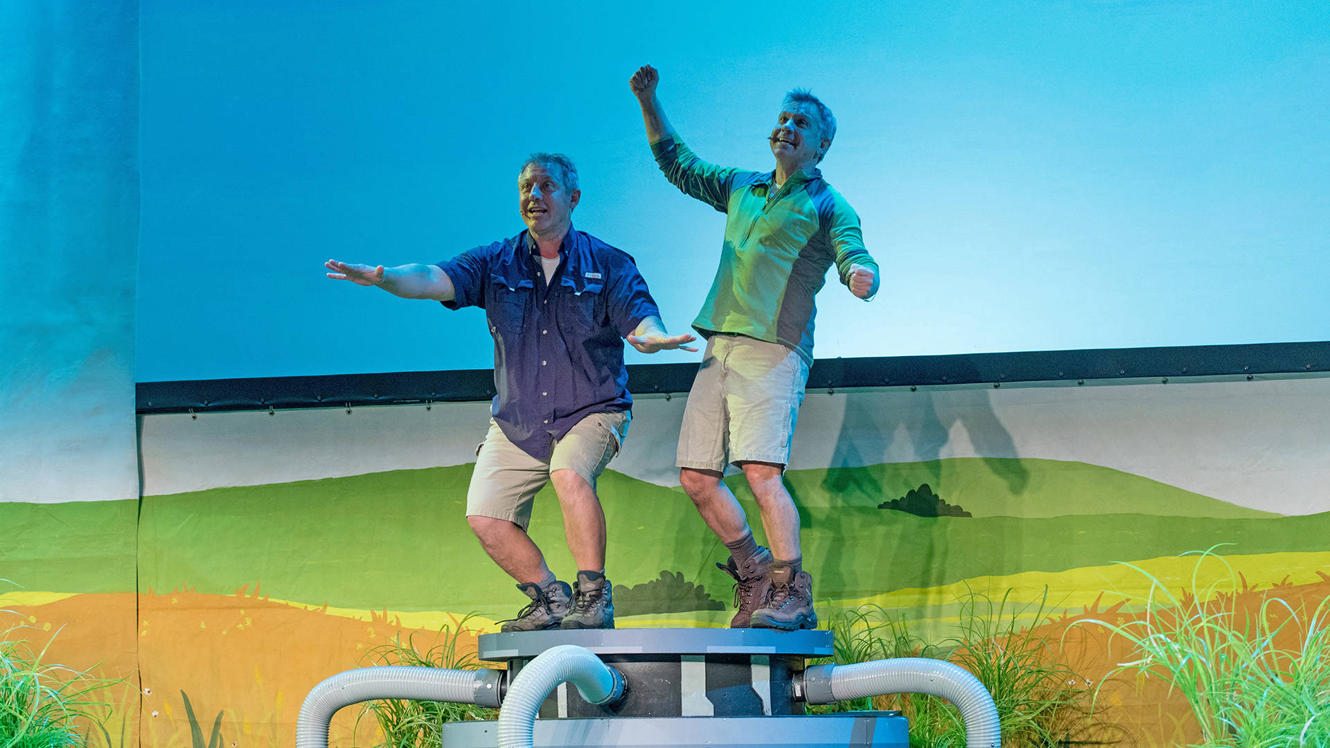 Kratt brothers, two men with light complexions wearing shorts and hiking boots, stand and cheer on top of a platform during a performance.