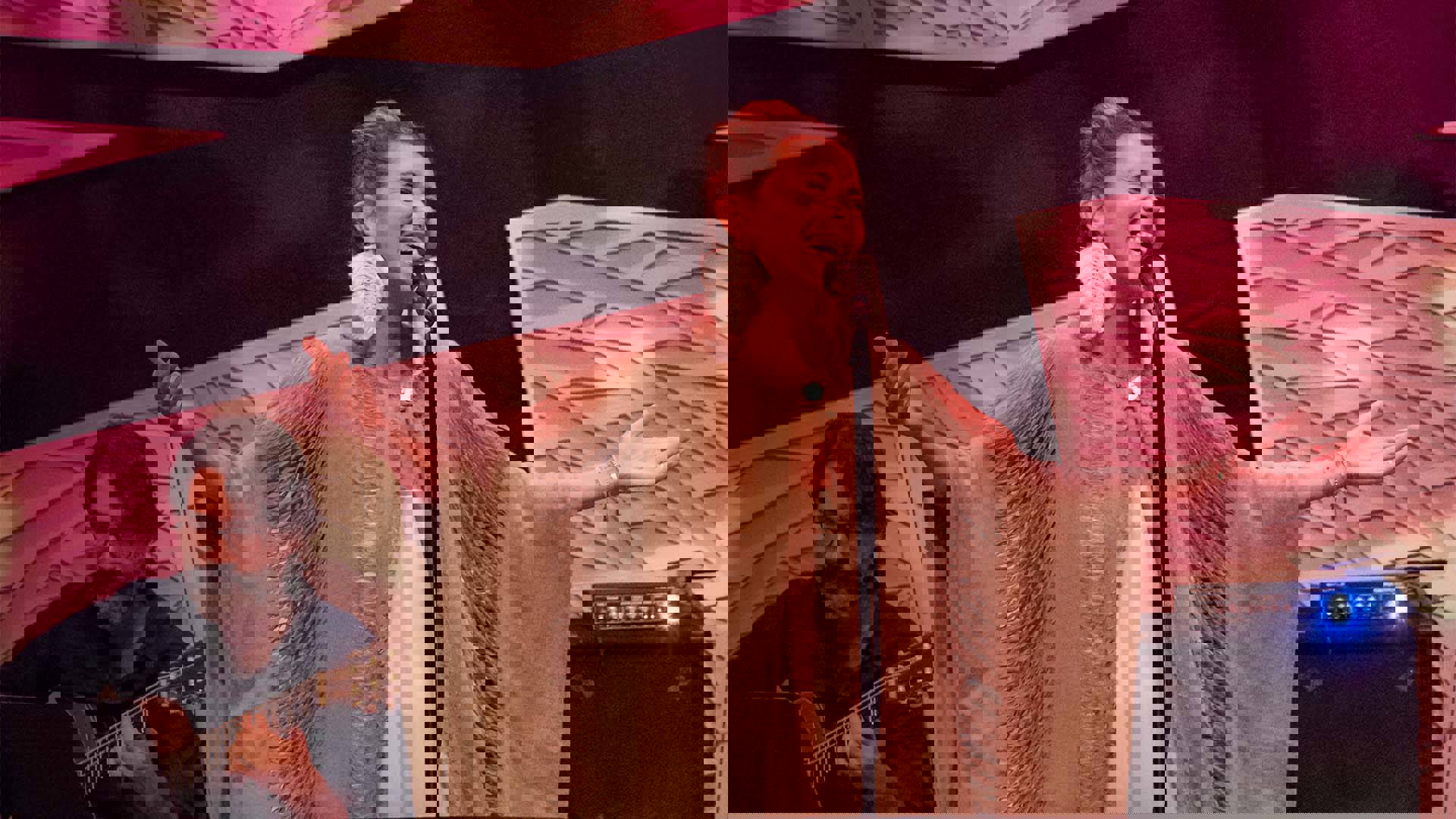 Magos Herrera, a Mexican woman with light brown long hair in a bun performing on stage. She is wearing large white feather earrings and an oversized brown fringed shawl.
