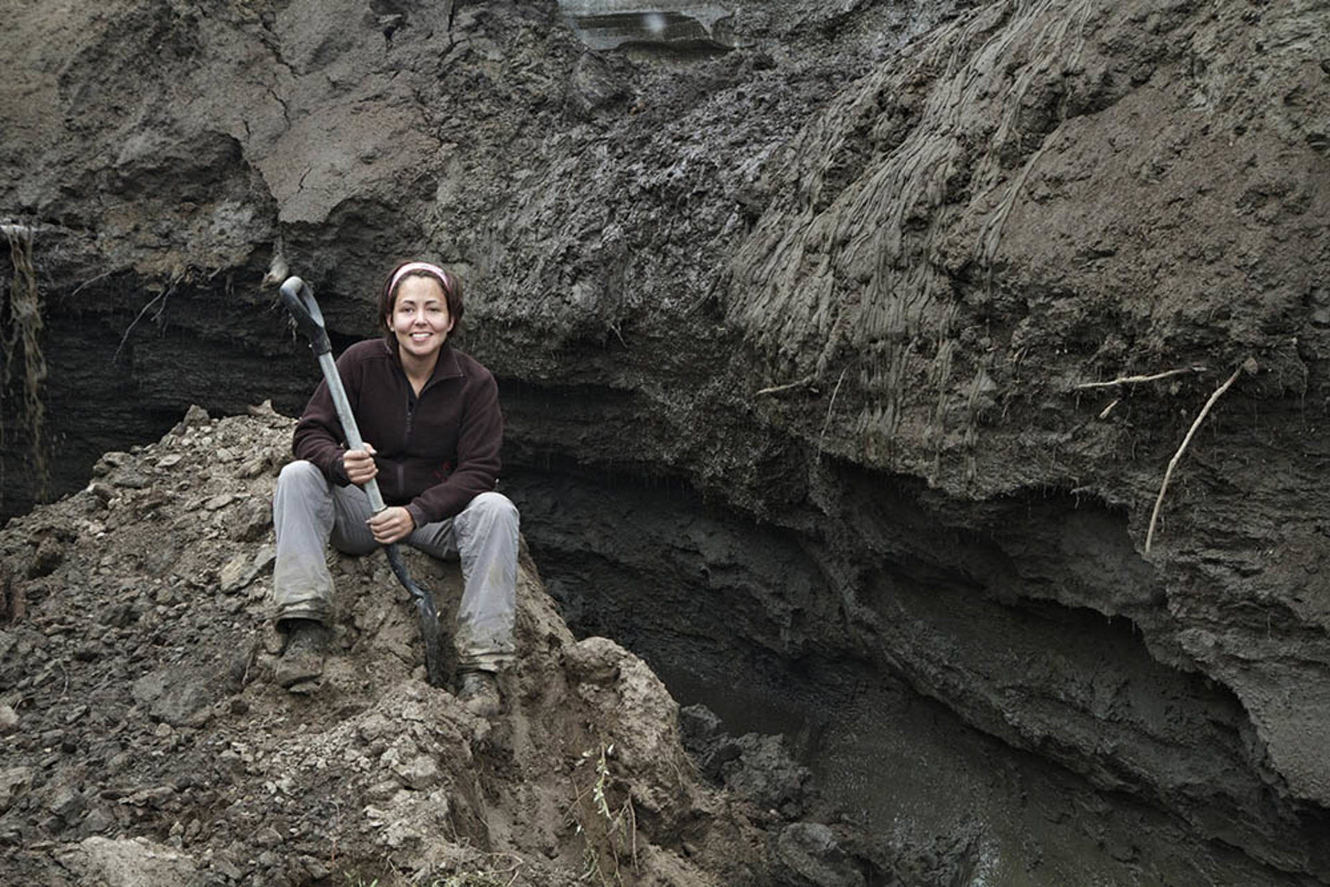 Beth Shapiro holding a shovel and sitting on a pile of dirt. Behind her is a muddy wall.