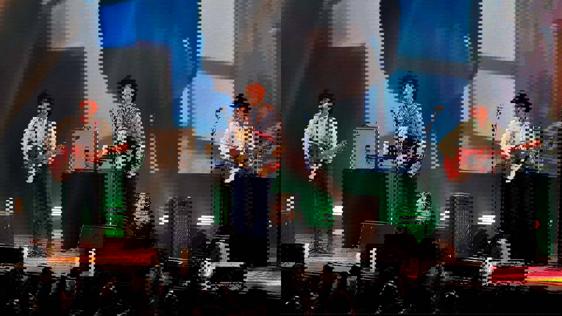 Two guitarists and the singer/accordion player performing on stage for Intocable. They are all wearing jeans and black cowboy hats with long sleeve collared shirts.