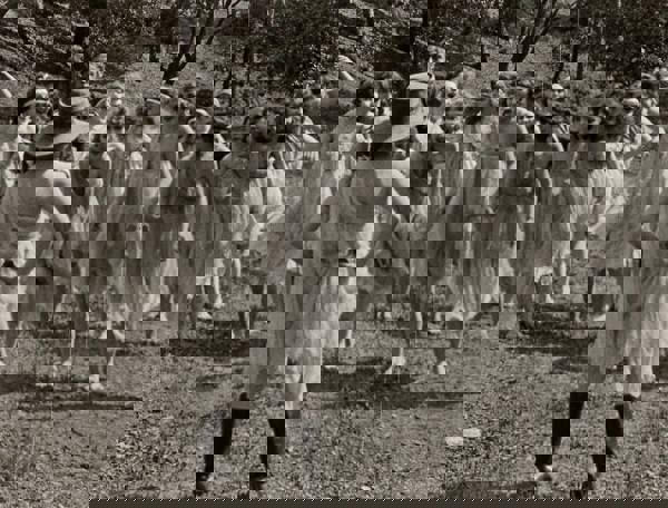 Vintage black and white image of a woman with light complexion in a white, torn dress and cowbody boots and hat, standing with her hands on her hips, facing a crowd of similarly dress women barefoot in a field.