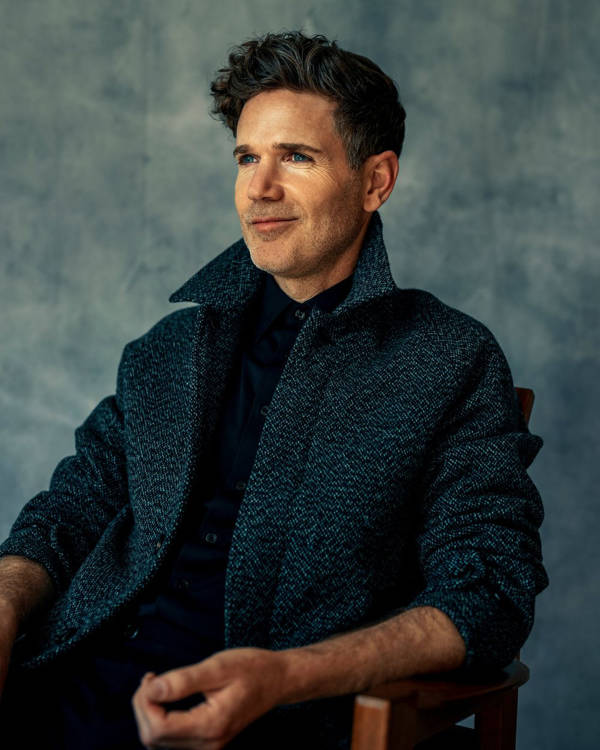 A young man with light complexion, short dark hair, a black shirt and grey jacket seated and looking off center.