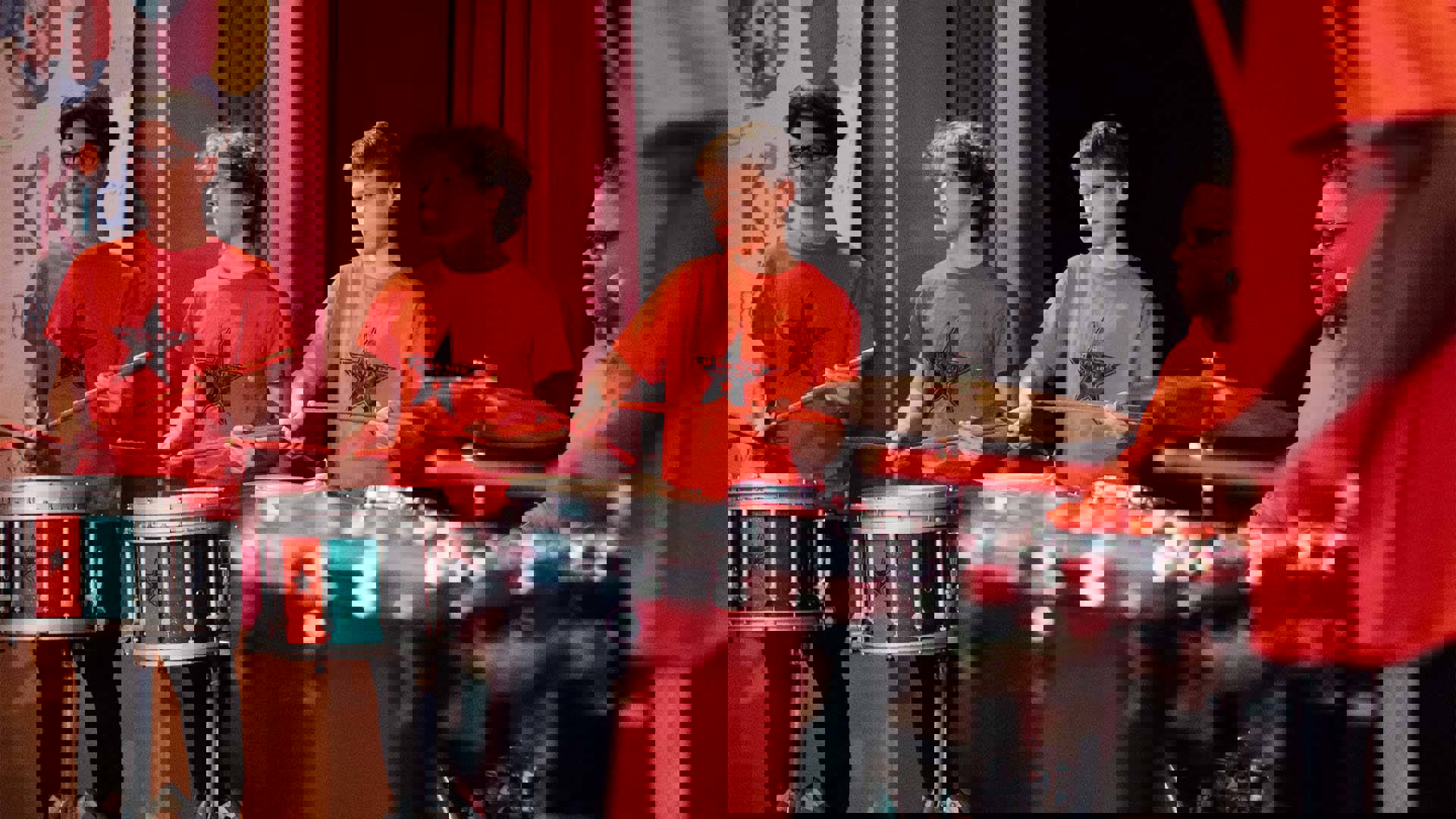 Children in orange shirts playing red green and black snare and tenor drums. A middle aged african american man with glasses and a beard is playing a drum set.