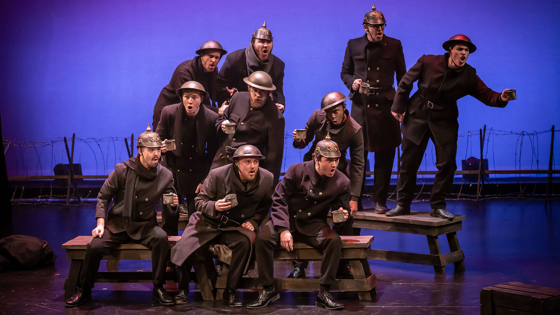 Ten actors in WW1 soldiers outfits sing a song against a blue background. They are sitting and standing on wooden benches.