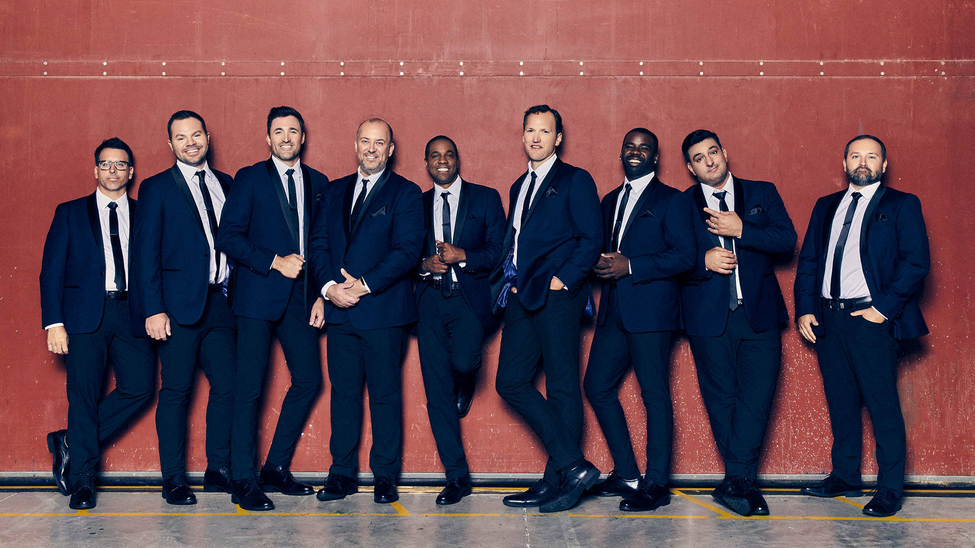 Nine men wearing blue suits standing in front of a red wall.