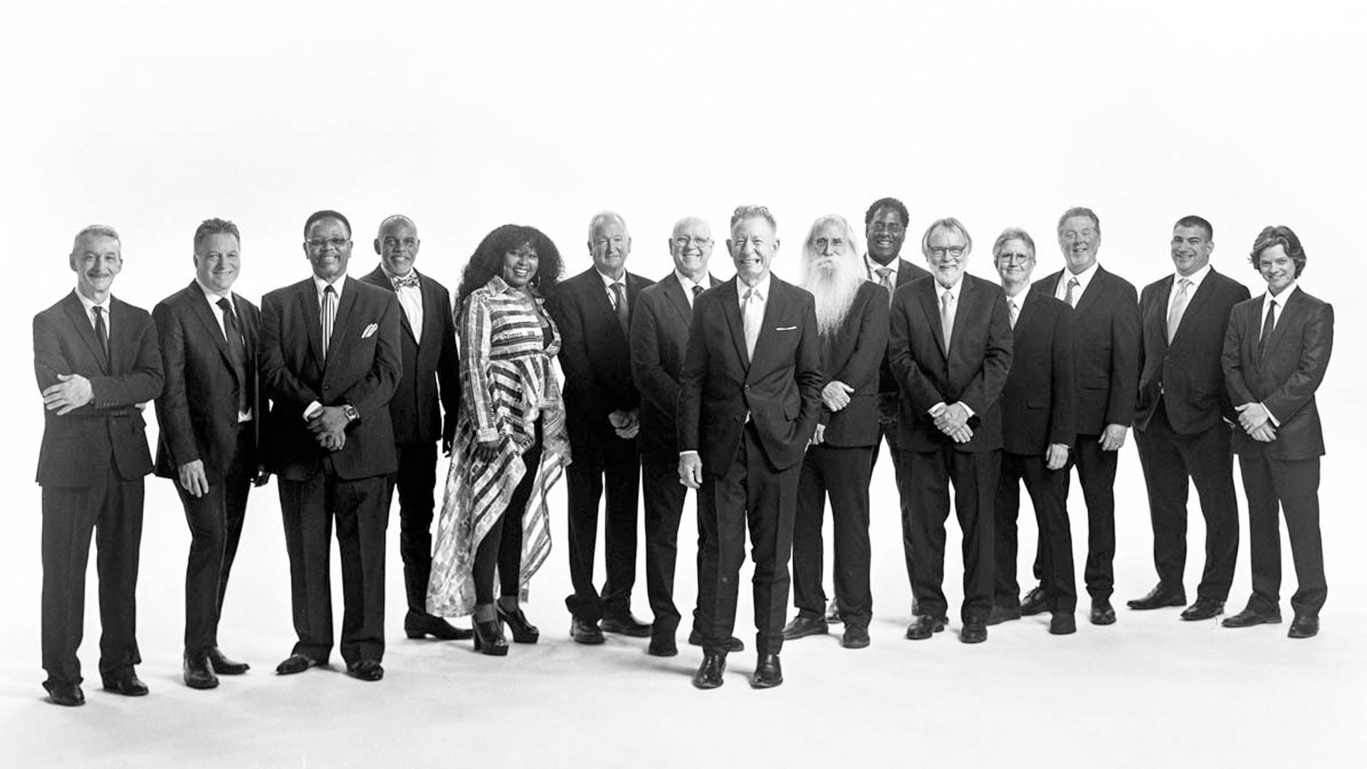 Fifteen people standing in a line together smiling at the camera in formal wear. At the center is Lyle Lovett, an older caucasian man with thinning short grey hair wearing a dark suit with a silver tie. Photo in black and white.