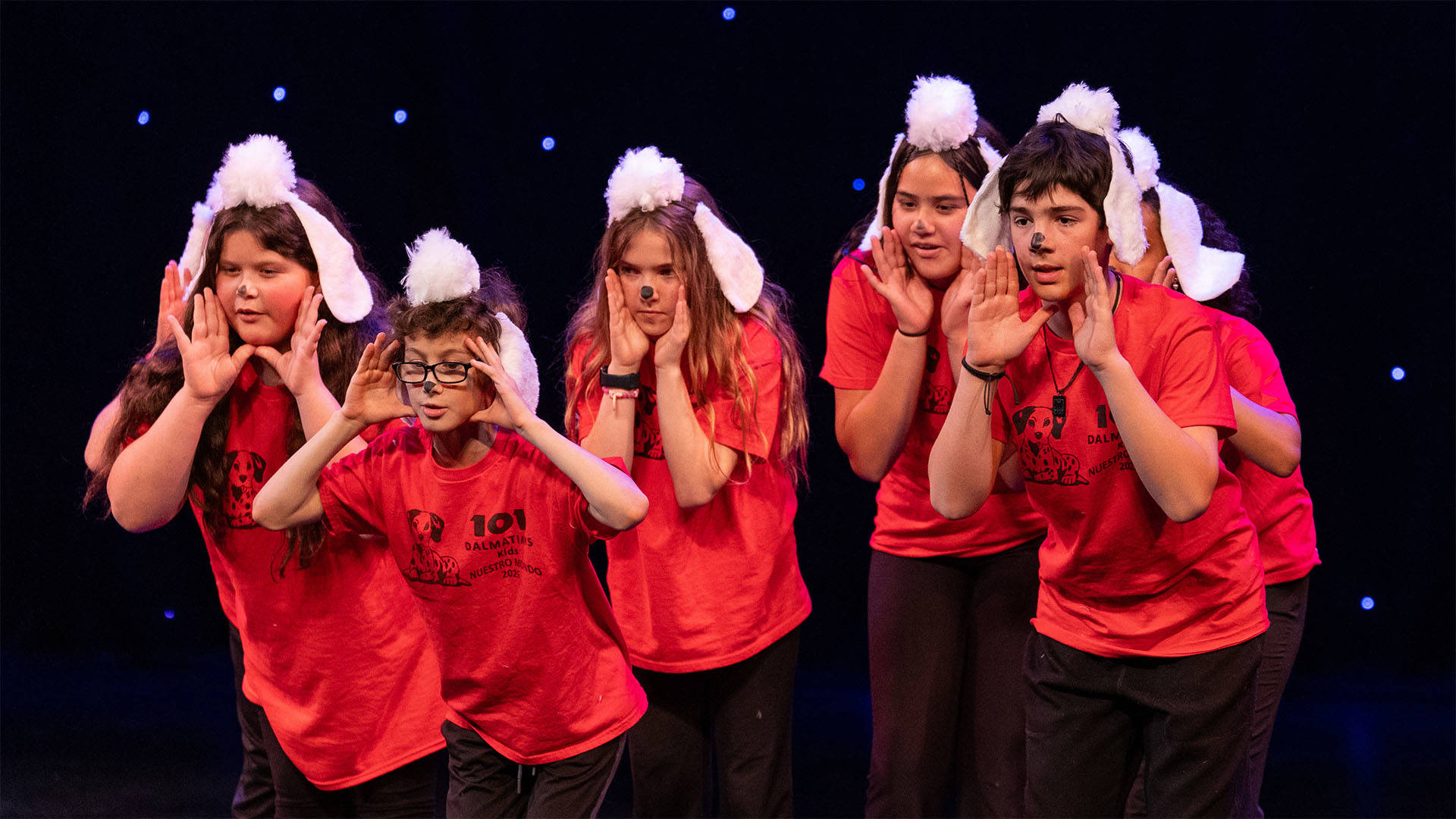 Young students in red shirts and puppy ear headwear performing on a dark stage. 