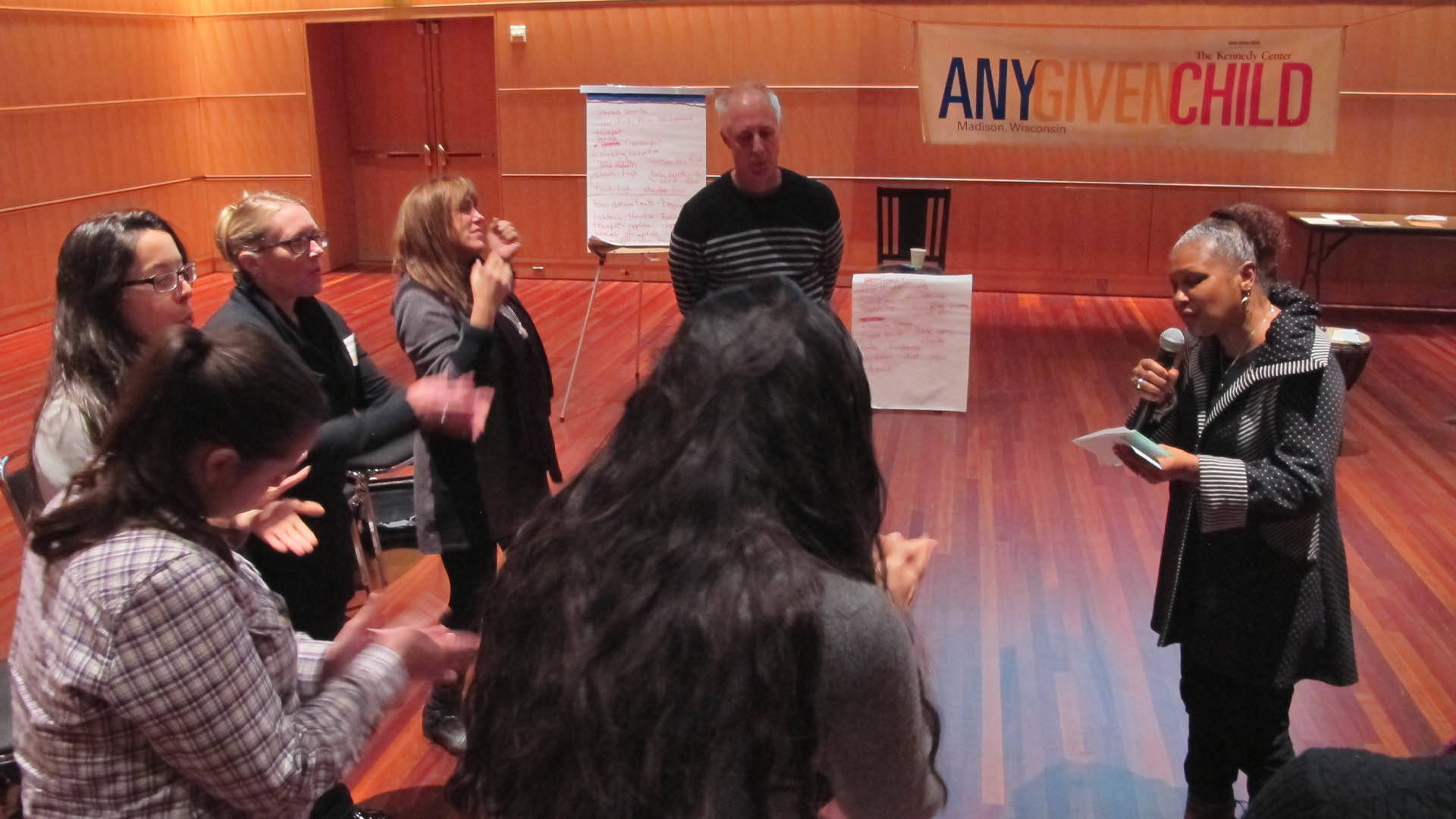 Several adults doing hand exercises in a circle. One woman stands with a microphone giving them instructions.