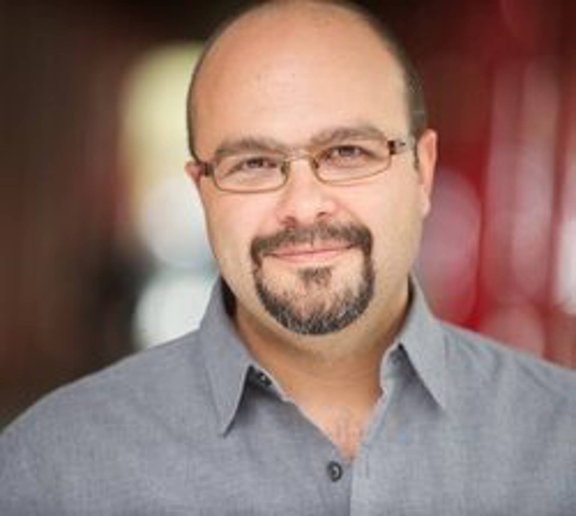 Headshot of a man with a bald head, goatee and glasses in a grey dress shirt.