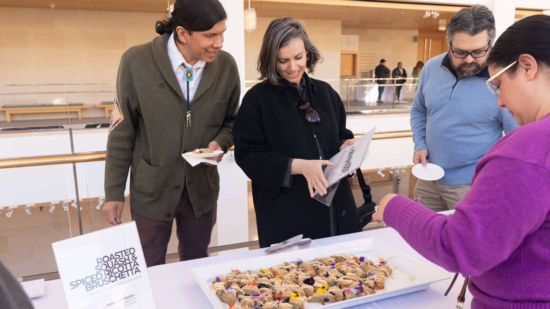 A group of people smile and look at a sign describing colorful baked goods that are on a platter below them.