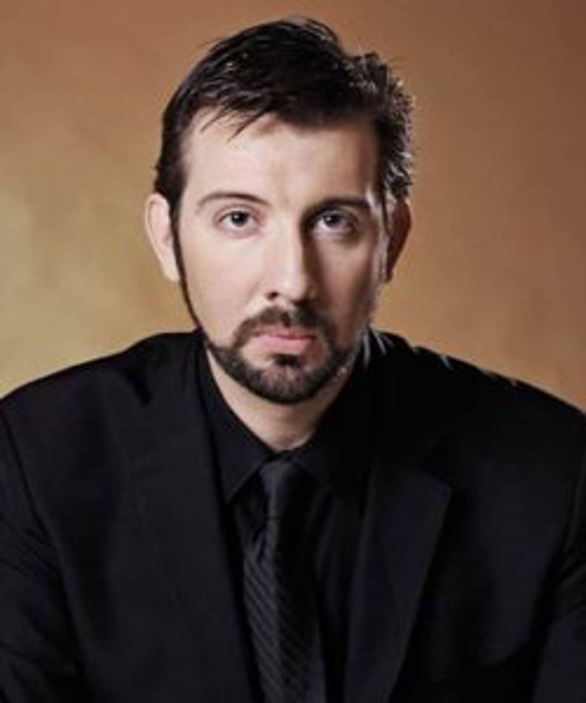 Headshot of a caucasian man with short dark hair and dark eyes in a black suit.