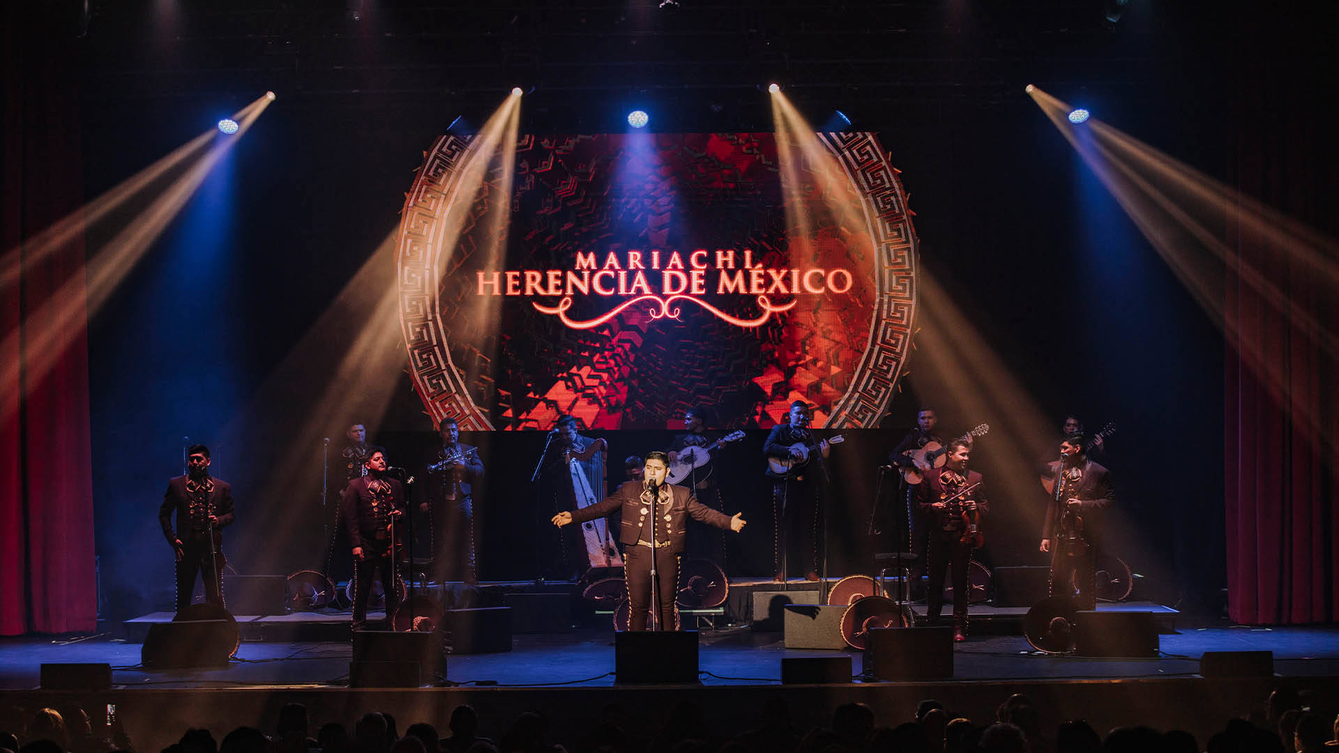 Two rows of people standing on a stage. There are five in the front row and seven on risers in the back row. They are wearing dark Mariachi outfits. Behind them is a screen that shows the words "Mariachi Herencia de Mexico".