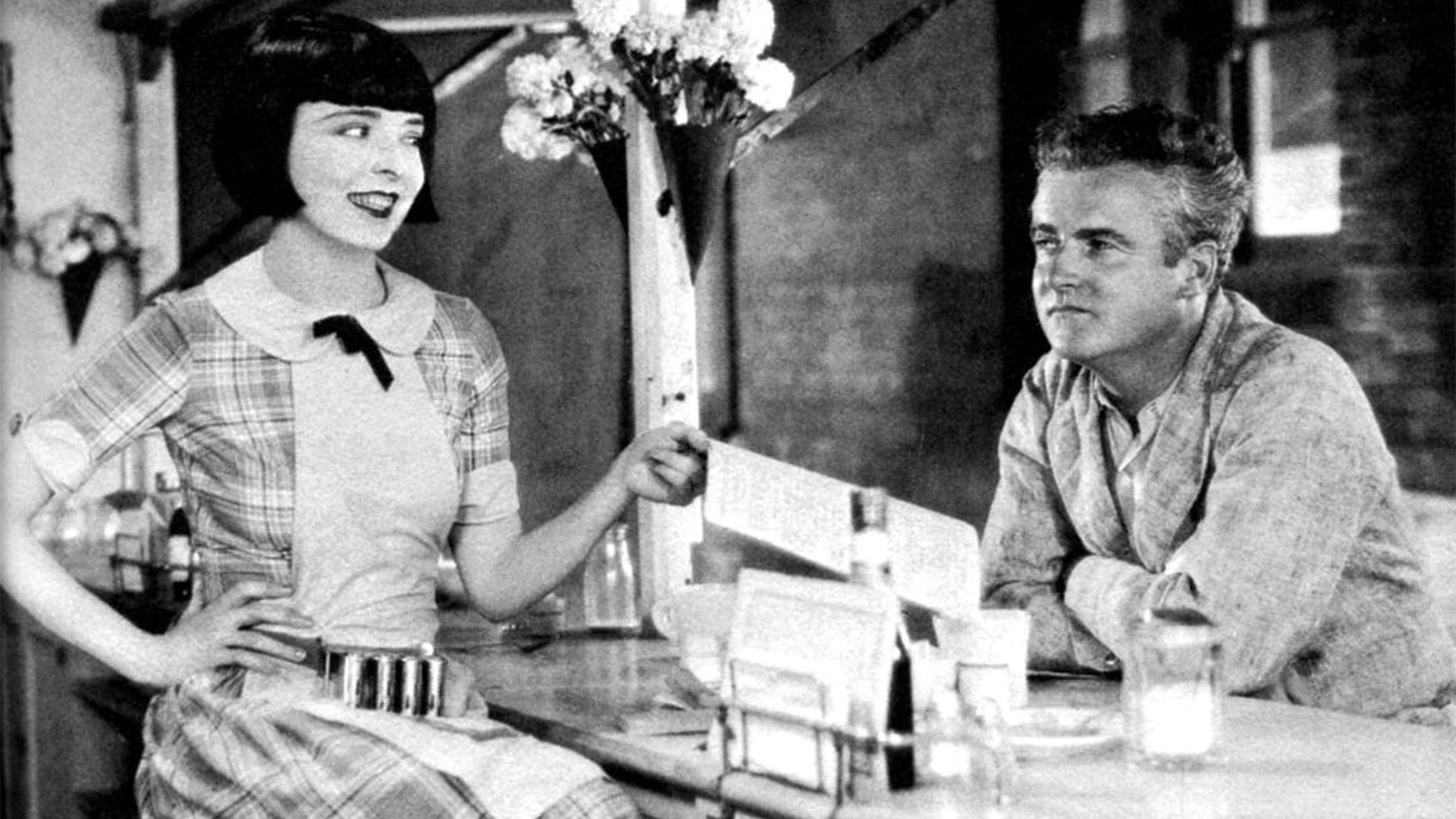 vintage black and white image of a light complexion woman with medium straight black hair sitting at a diner counter and smiling. She is holding a menu out for a light complexion man leaning on the counter with a look of suspicion