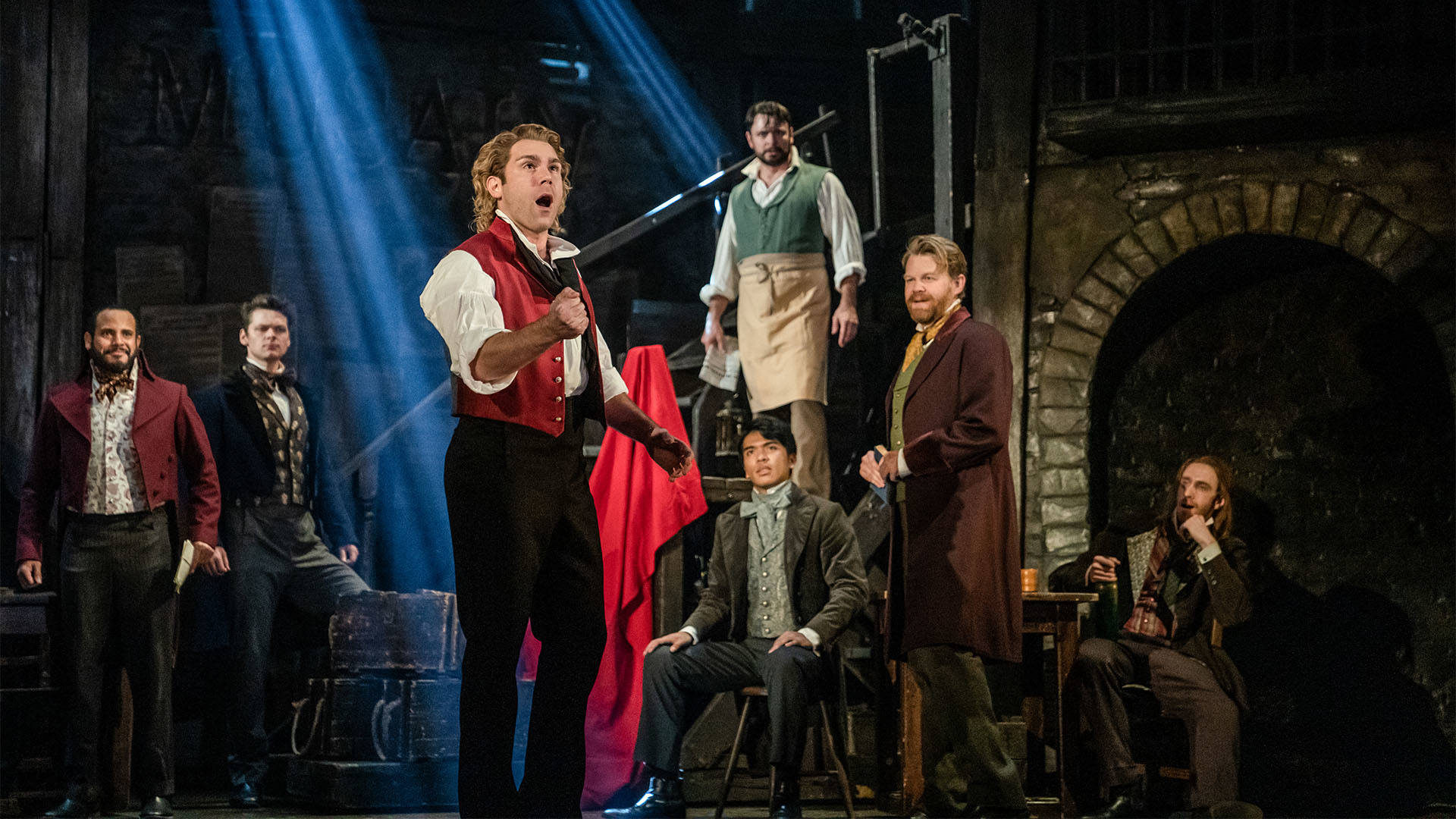 The cast of Les Miserables performing. At the center a man with long blond hair, a striking red vest and clenched fists sings.