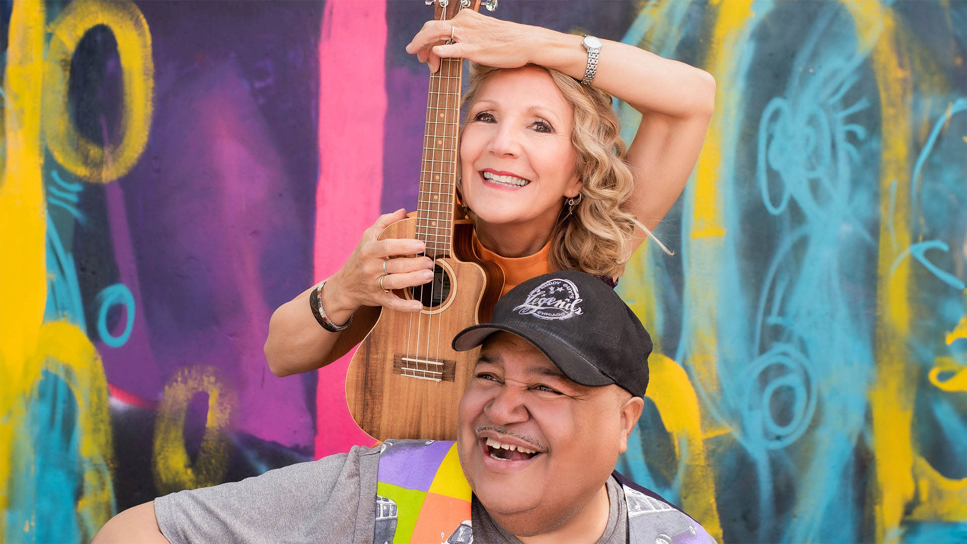 A smiling man and woman. The woman has light complexion and wavy blonde hair. She is holding a ukulele over the shoulder of the man, who has medium complexion and a black baseball cap. The background is colorful.