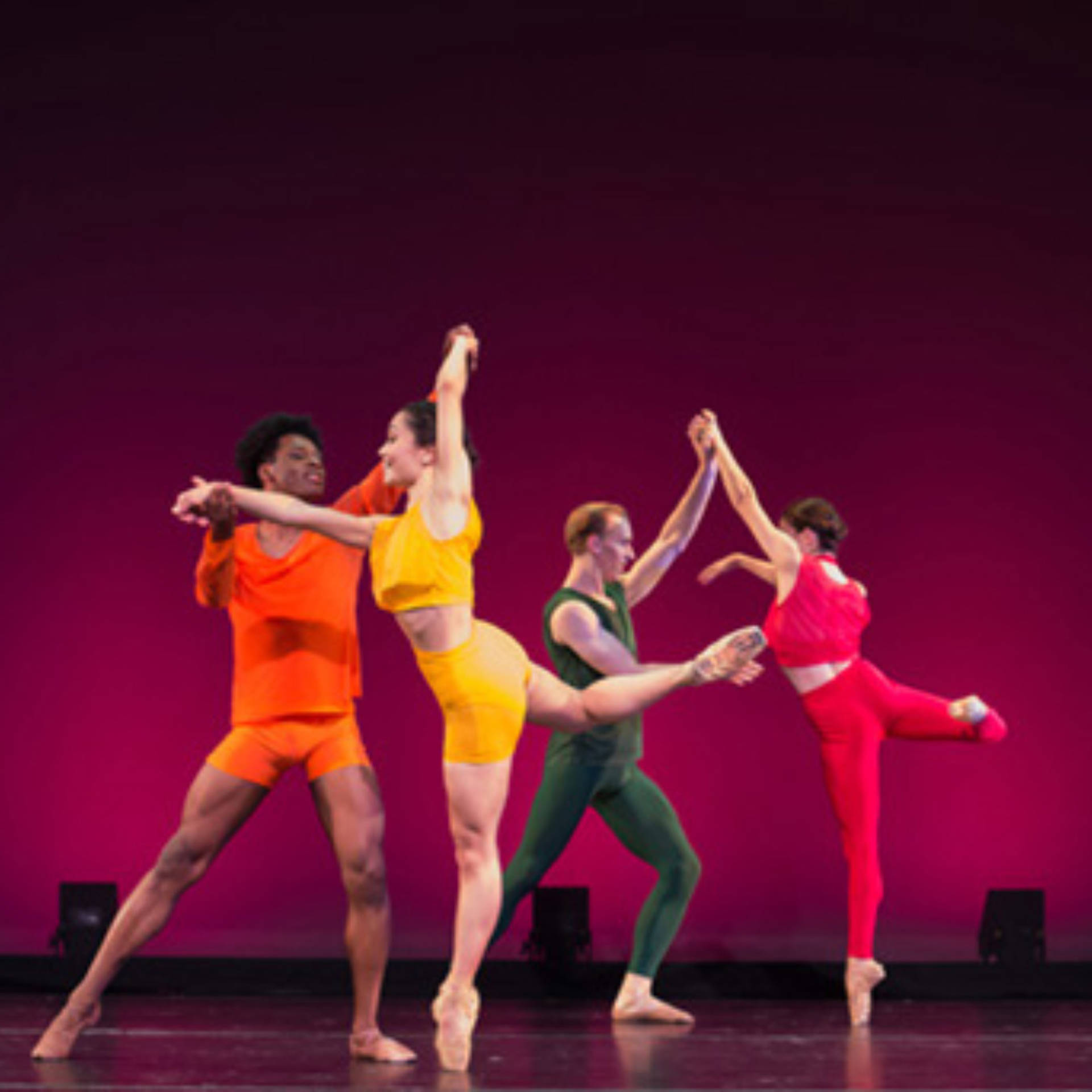 Four dancers in colorful clothing performing on a stage with maroon background.
