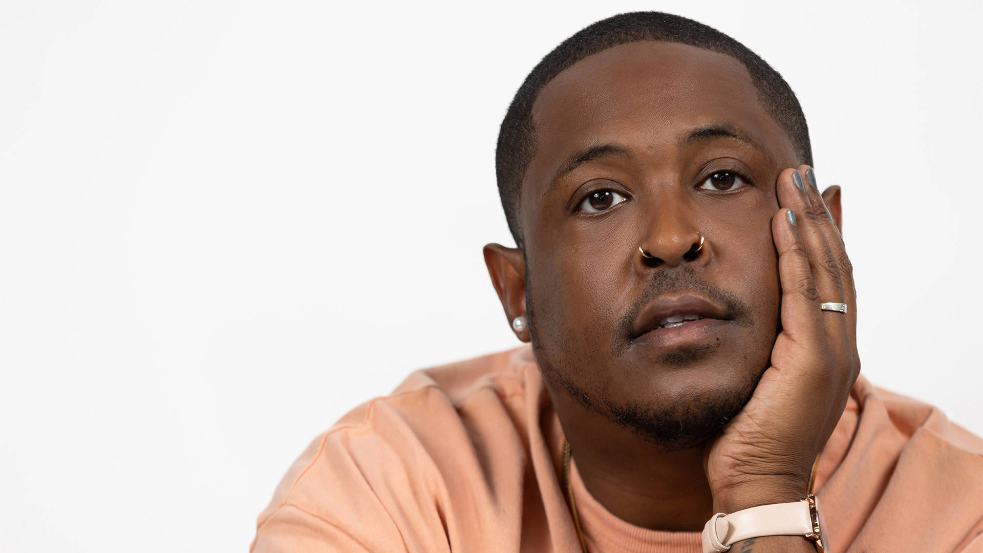A young african american man with short dark hair, brown eyes, and light facial hair looking towards the viewer with his head resting on his hand. He has a light orange shirt on and a creme colored watch.