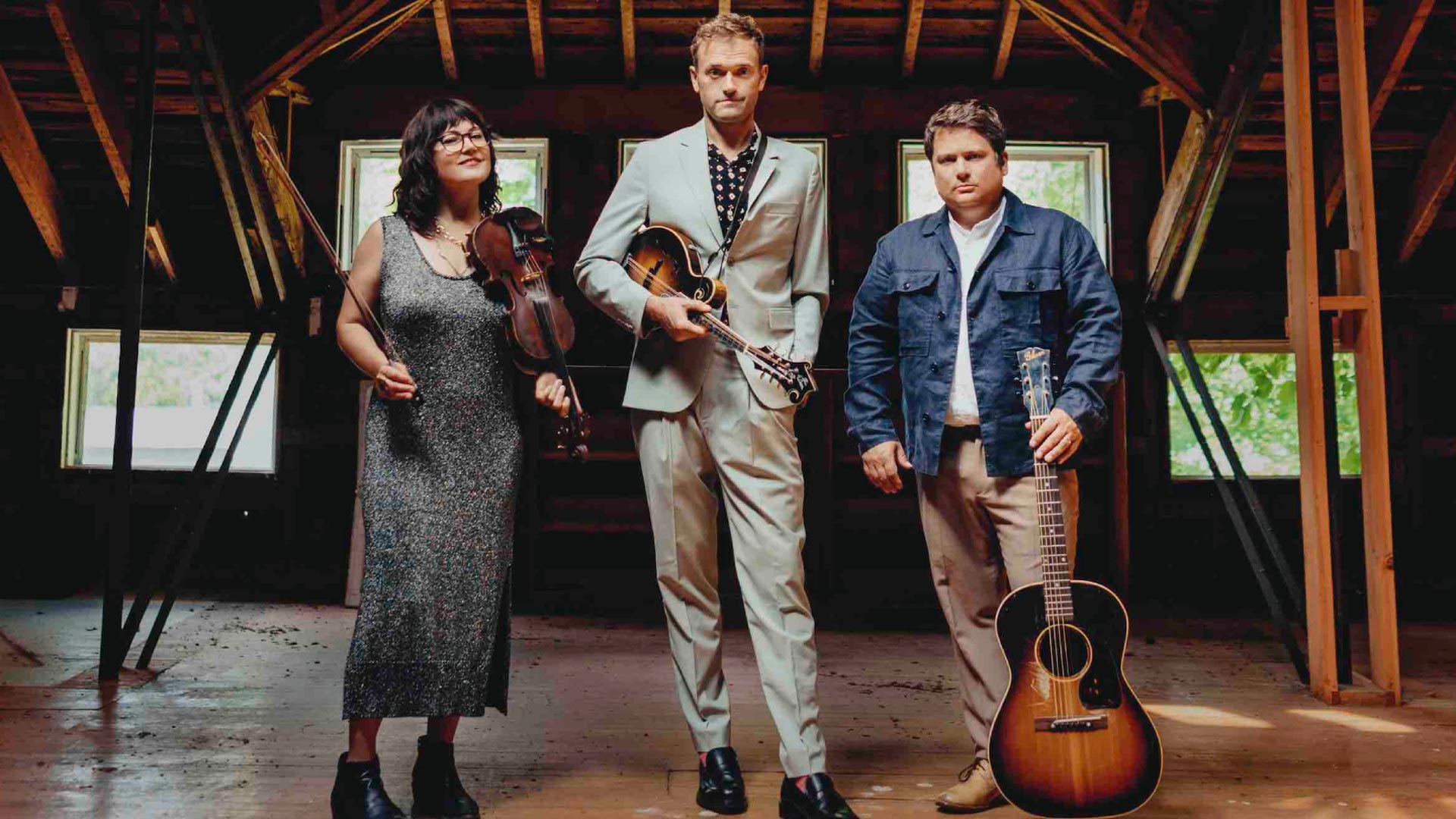 A bluegrass trio is standing in a barren room with wooden features and beams. A woman on the left has light complexion, brown hair and is holding a violin. The man in the middle has light complexion, blond hair, a grey suit and a mandolin. The man on the right has light complexion, brownish hair, a jacket and an acoustic guitar.
