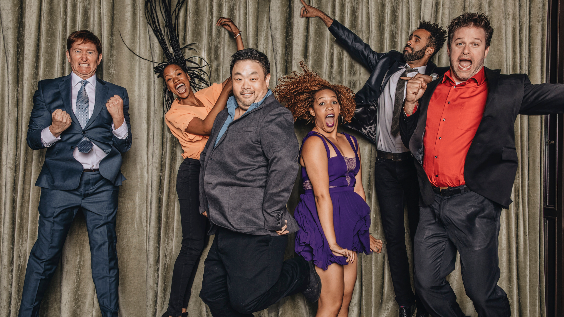 Six people jumping for a photo in front of a beige stage curtain. They are all wearing formal business clothes with mouths open in shock, surprise and silliness.