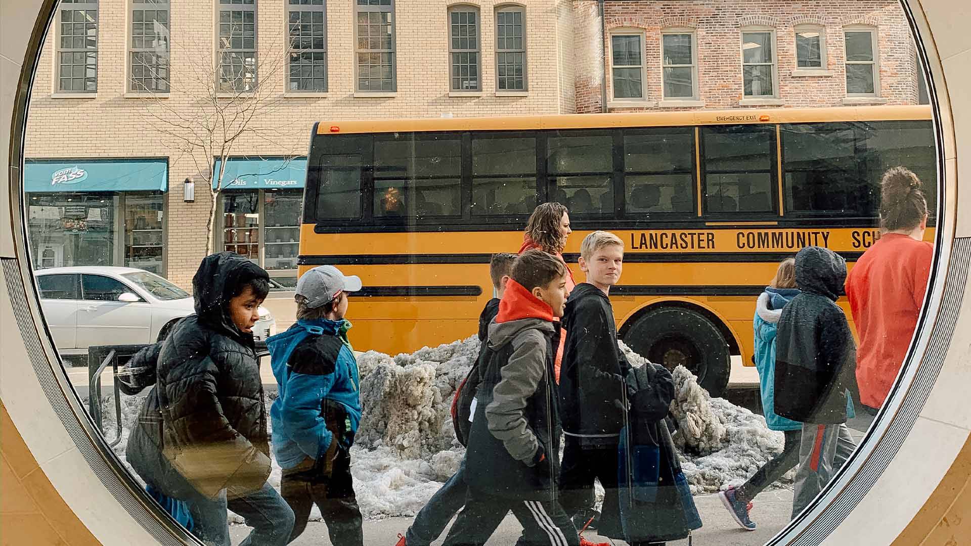 Several students walking past a round window outside of Overture. A school bus passes by in the background.